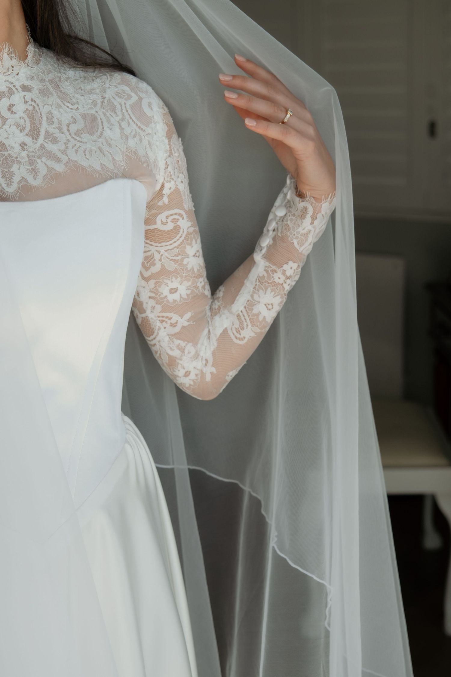 Bride in lace long-sleeve wedding dress holding flowing white veil indoors.