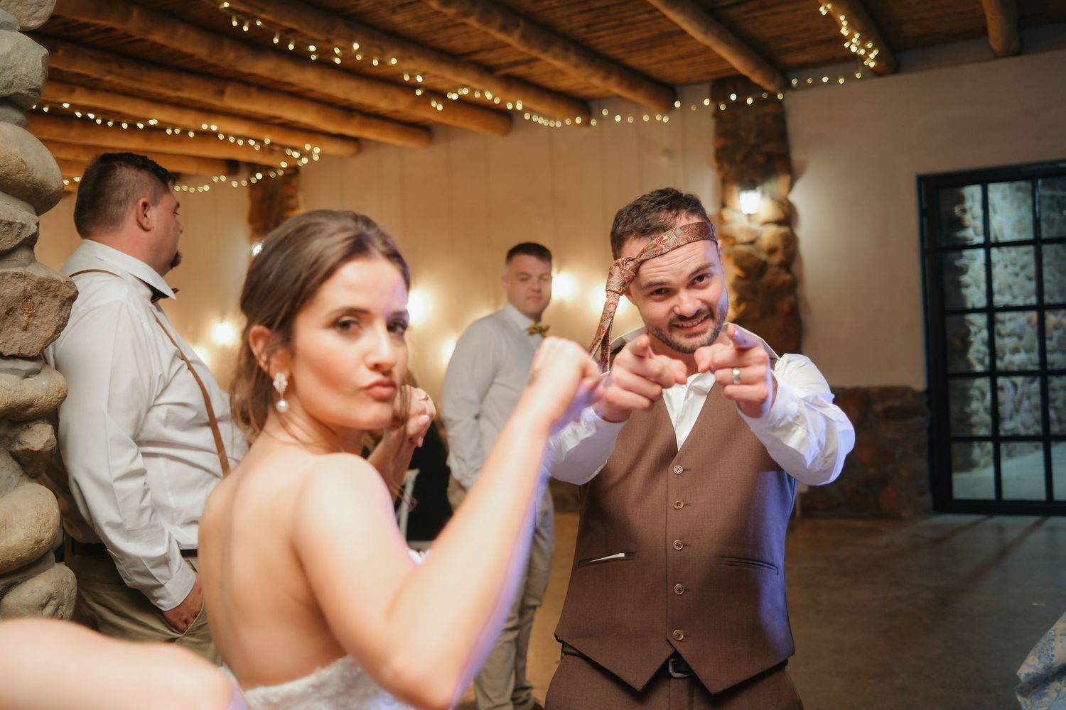Bride and groom playfully posing together on dance floor during lively wedding reception under string lights.