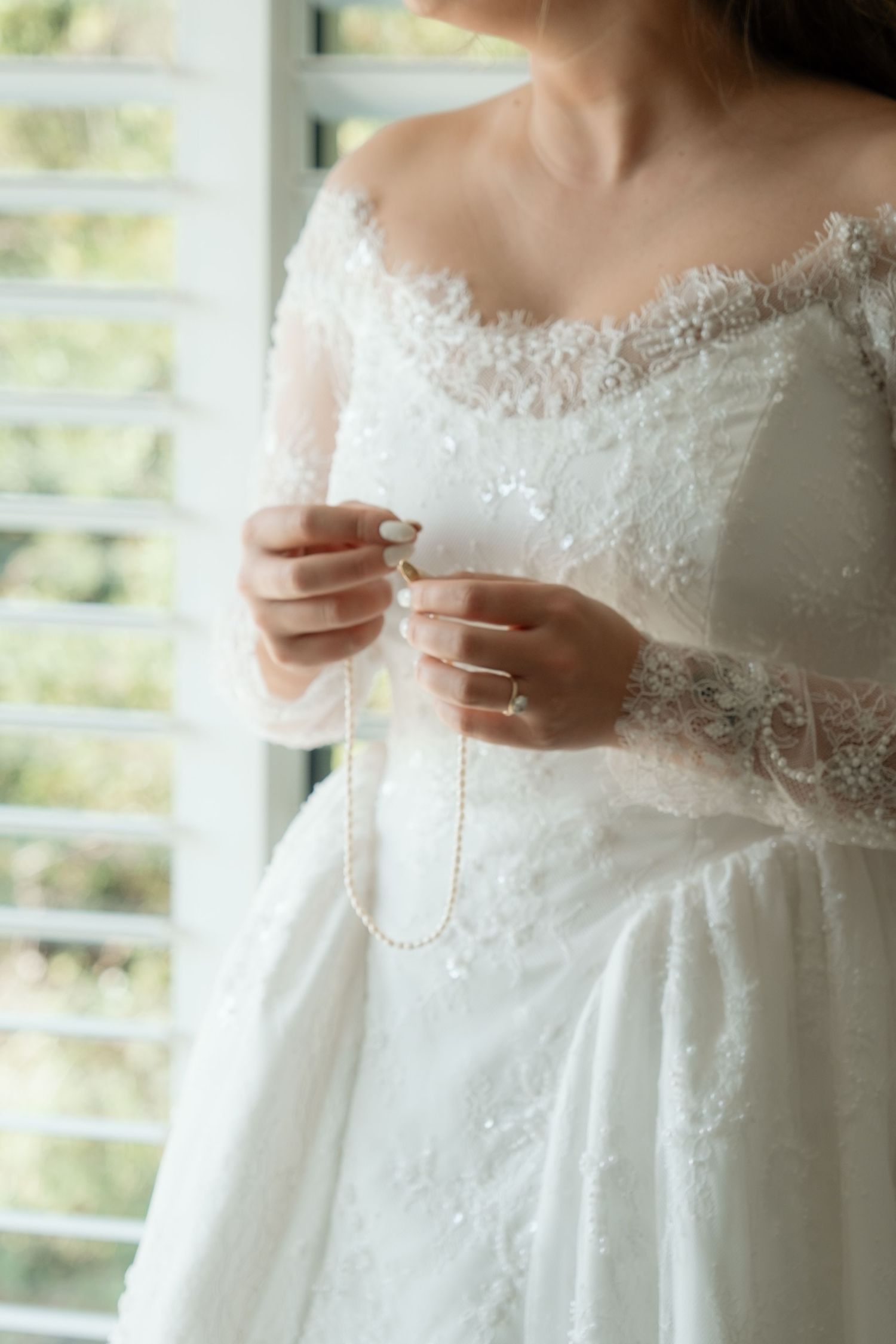Bride in off-shoulder lace wedding gown holding delicate pearl necklace by window.