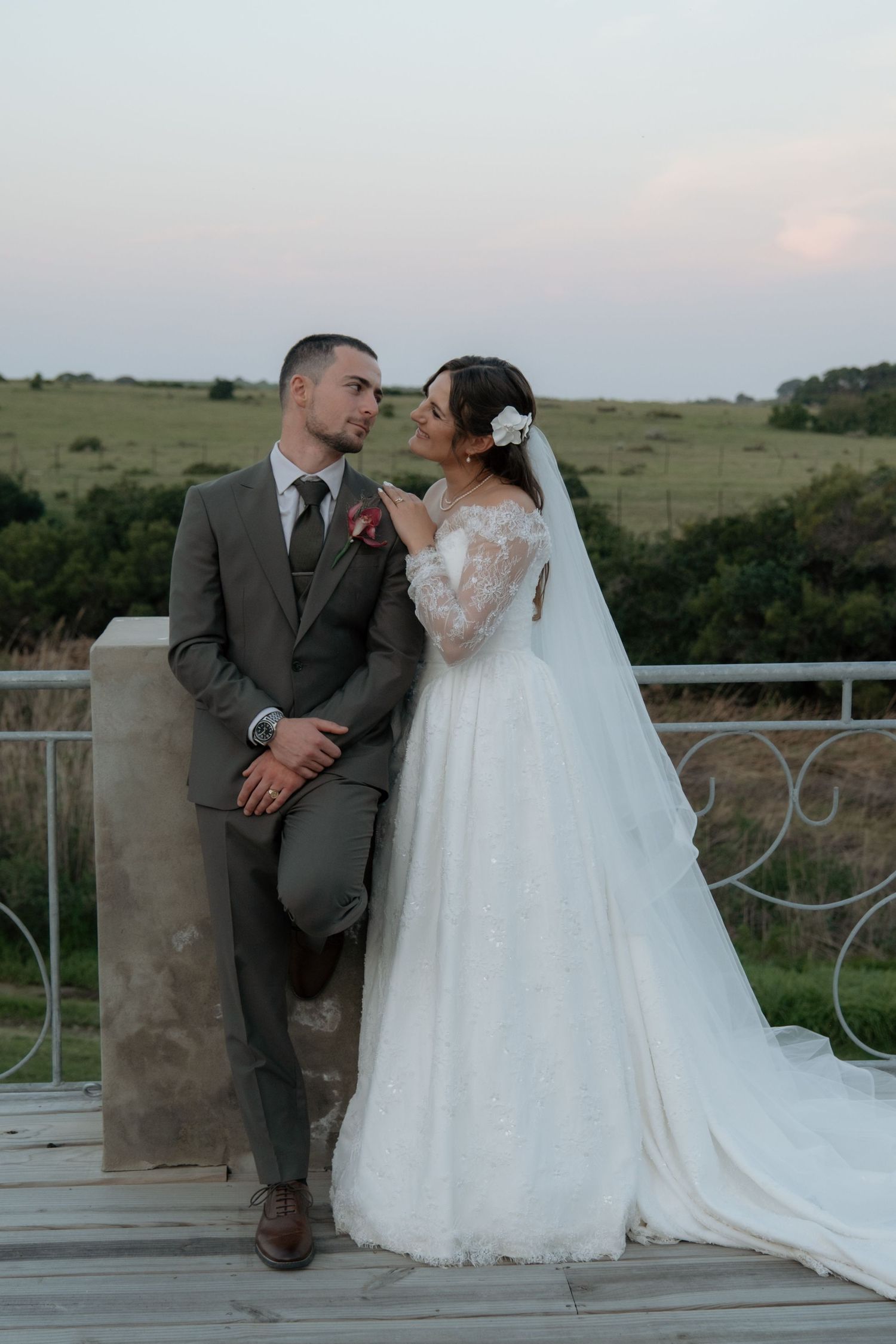 A bride and groom share a romantic moment on a terrace, with lush green fields and a pastel sunset sky behind them.