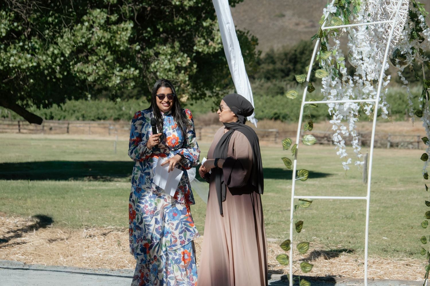 Two elegantly dressed women converse near a floral wedding arch on a sunny outdoor lawn.