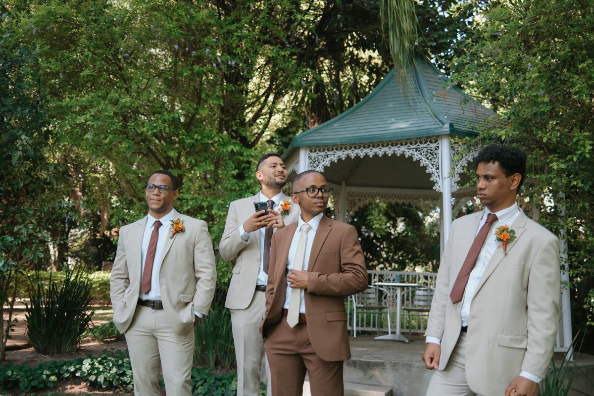 Groomsmen in beige and brown suits with burgundy ties gather near a white gazebo in a garden setting.