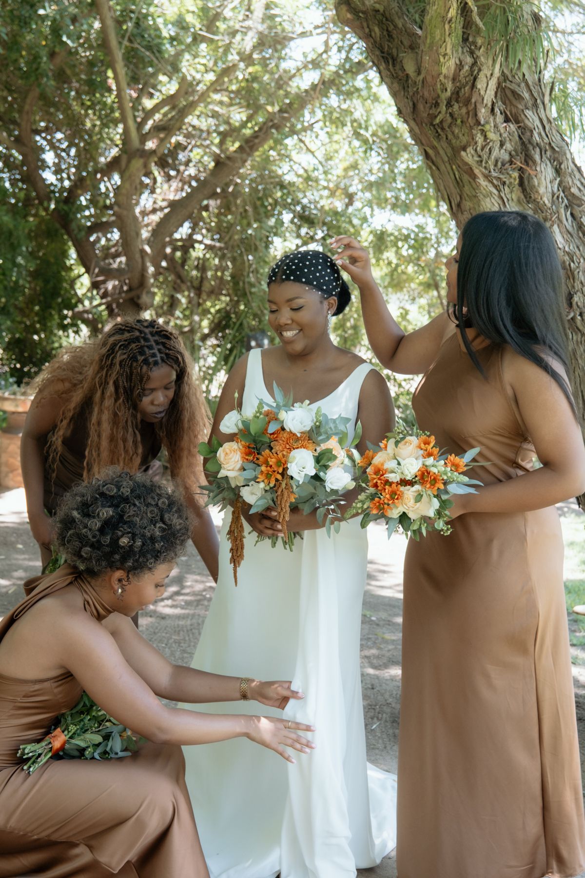 Bridesmaids in brown dresses help adjust bride's white gown while holding orange and white floral bouquets outdoors.