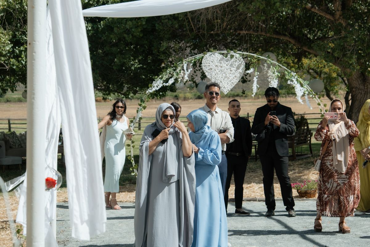 Wedding guests in formal attire walking under white draped outdoor ceremony arch on sunny day.