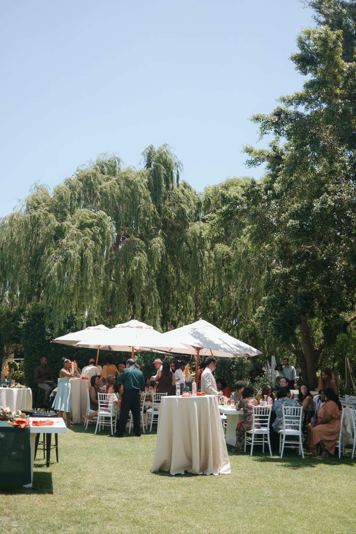 Outdoor wedding reception on green lawn with white tables, umbrellas, and guests mingling under large willow trees.