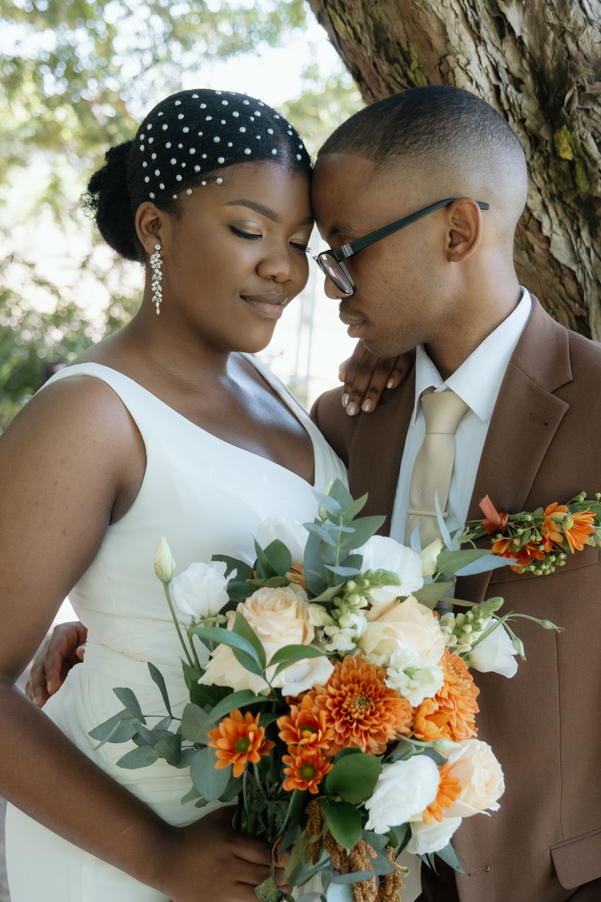 A Black bride and groom lean together tenderly by a tree, she holding a bouquet of orange and white flowers on their wedding day.