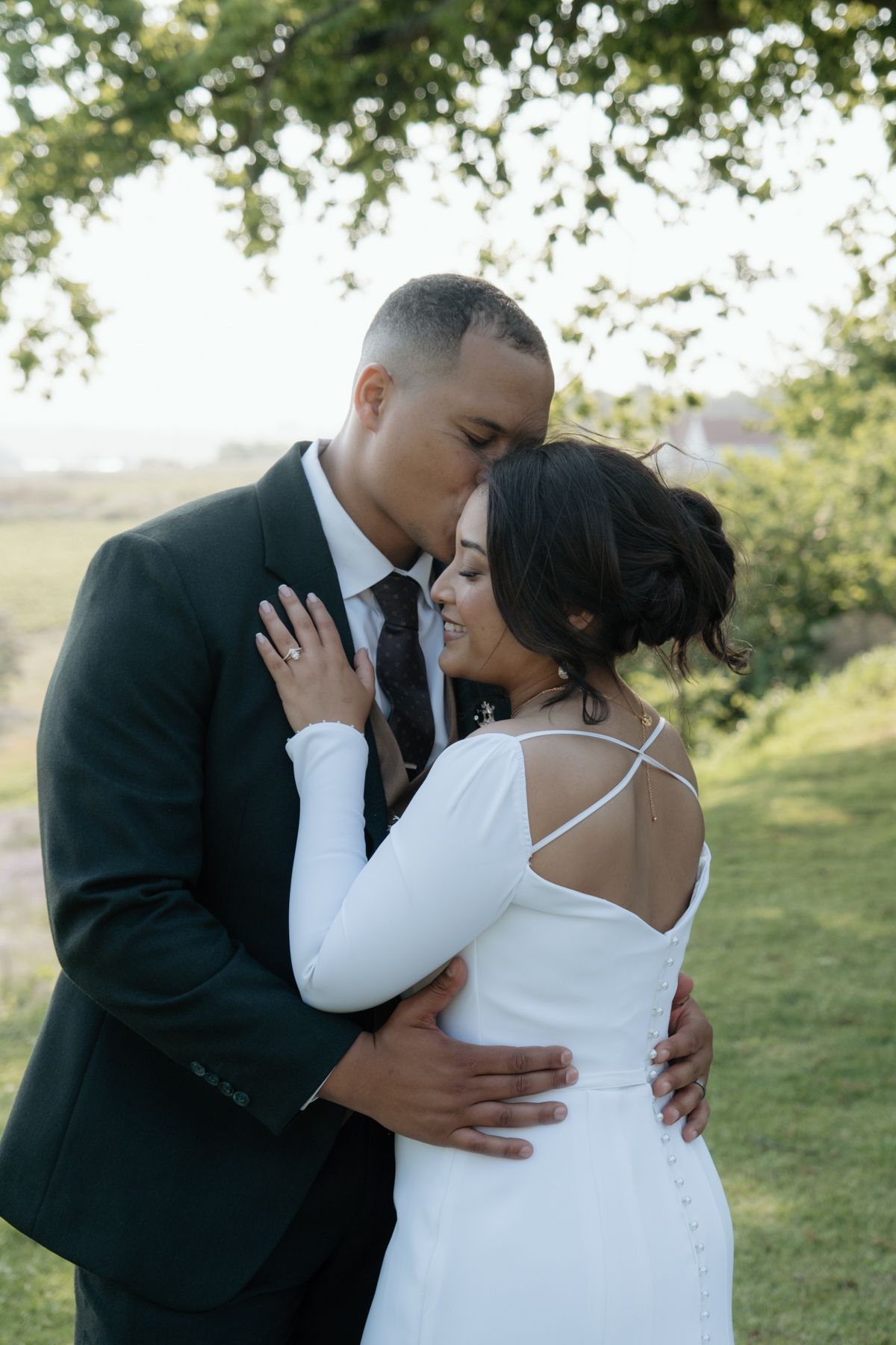 Groom in dark suit kissing bride in white spaghetti strap gown in a lush green outdoor setting.