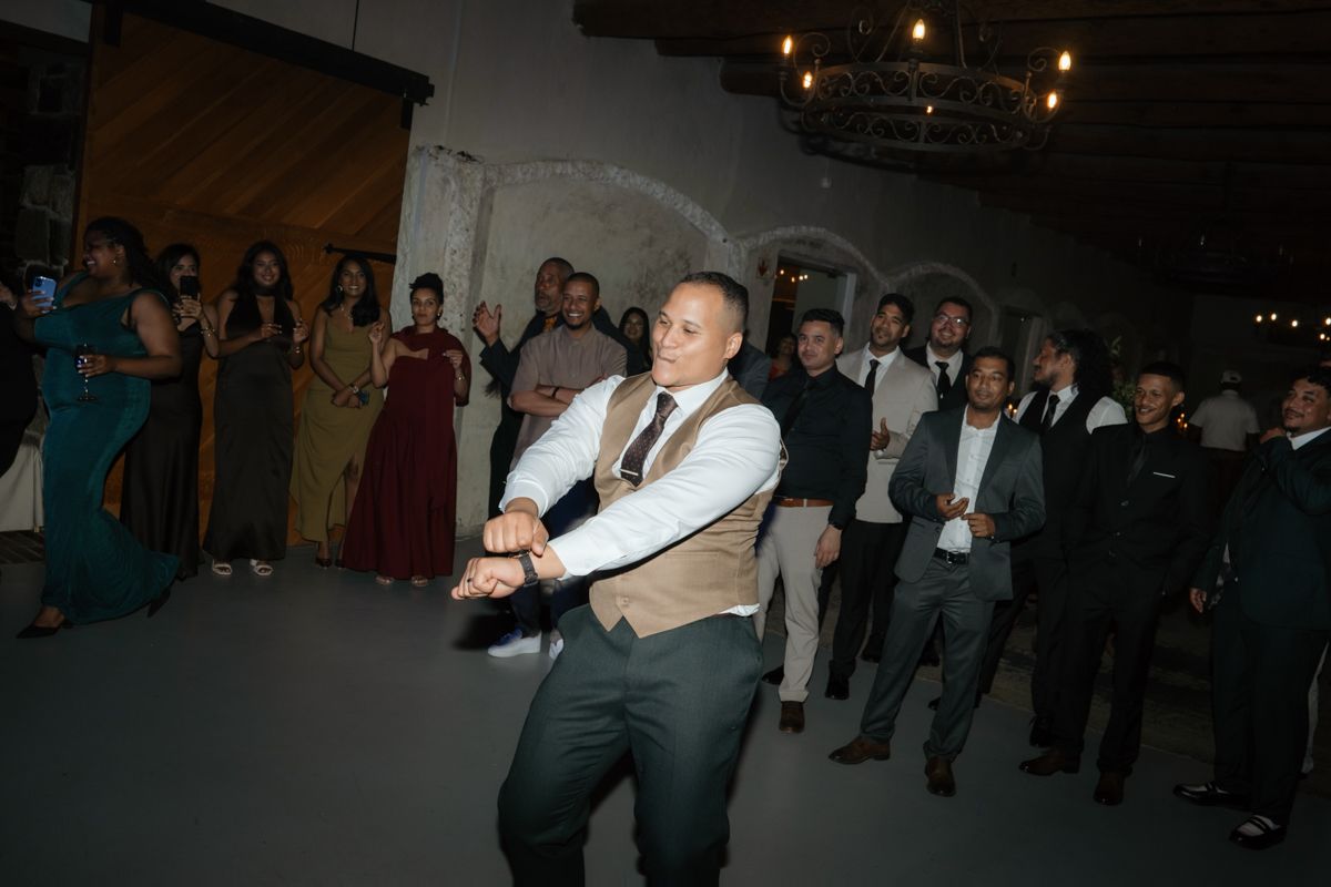 Groom in vest and dress shirt energetically dances at wedding reception surrounded by guests.