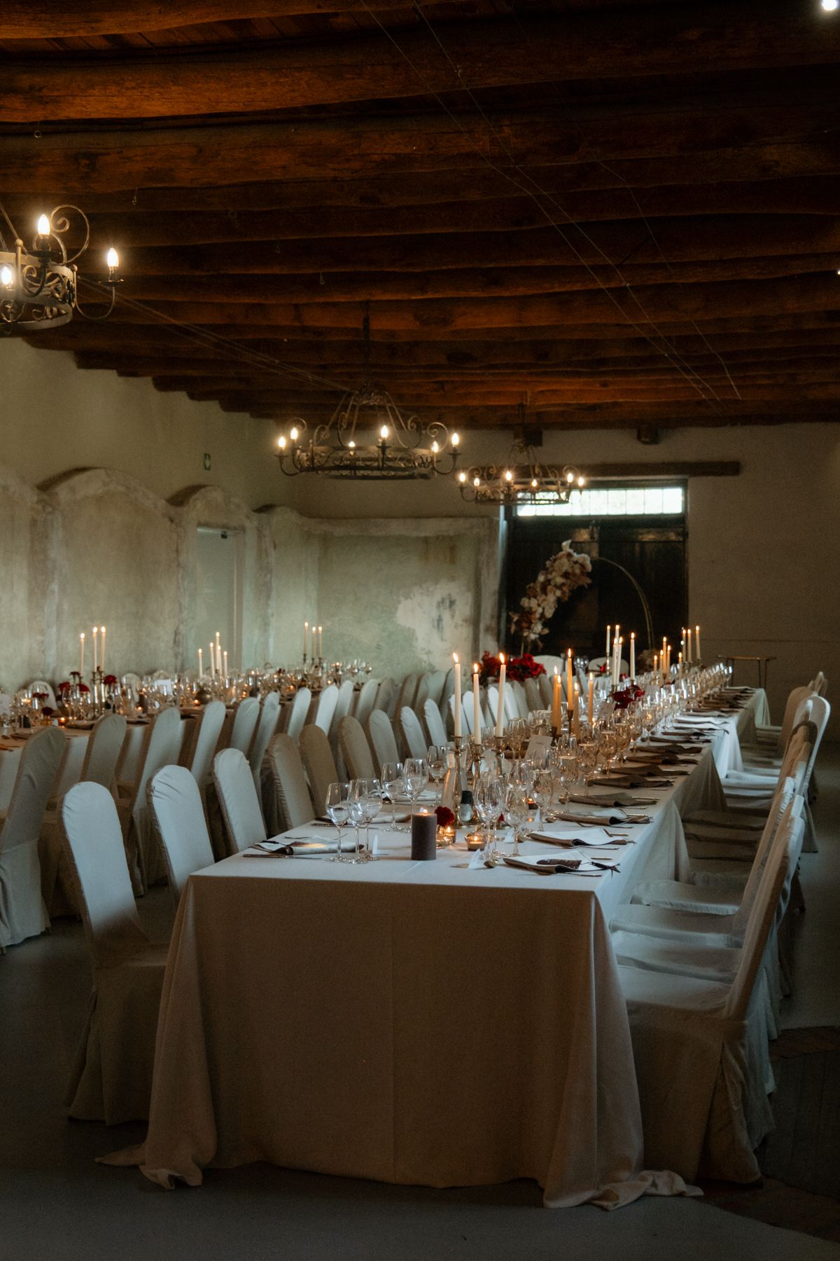 Elegant wedding reception hall with long tables, candles, white chairs, and rustic wooden ceiling.