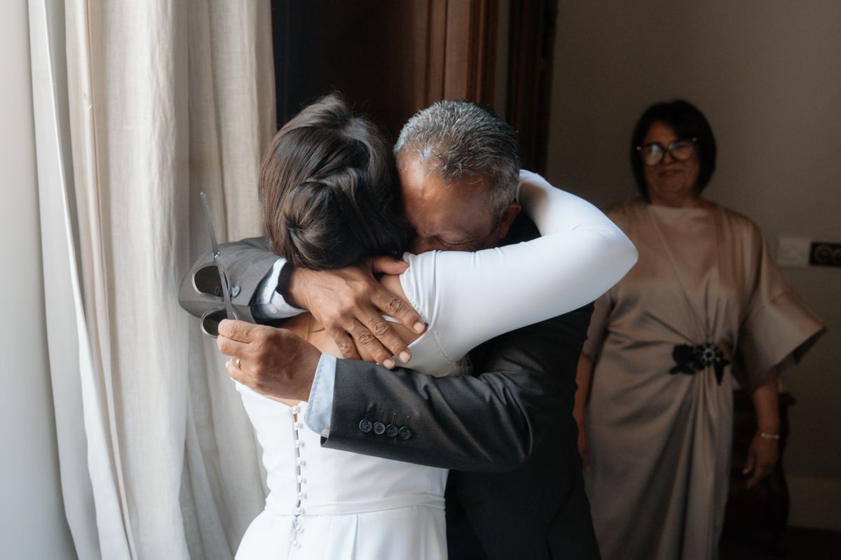 Emotional moment as bride in white gown receives a warm hug from older man at wedding venue.
