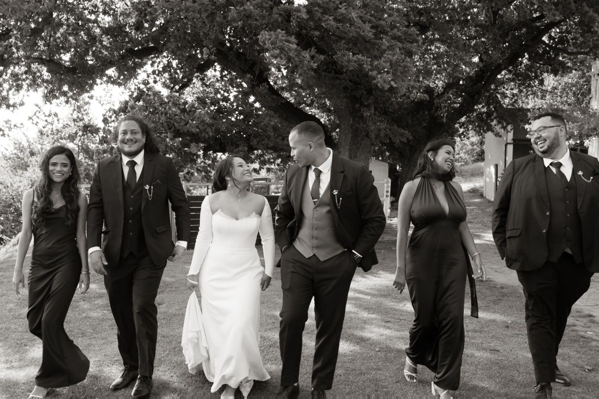 Black and white photo of bride, groom and wedding party walking together under a large tree outdoors.