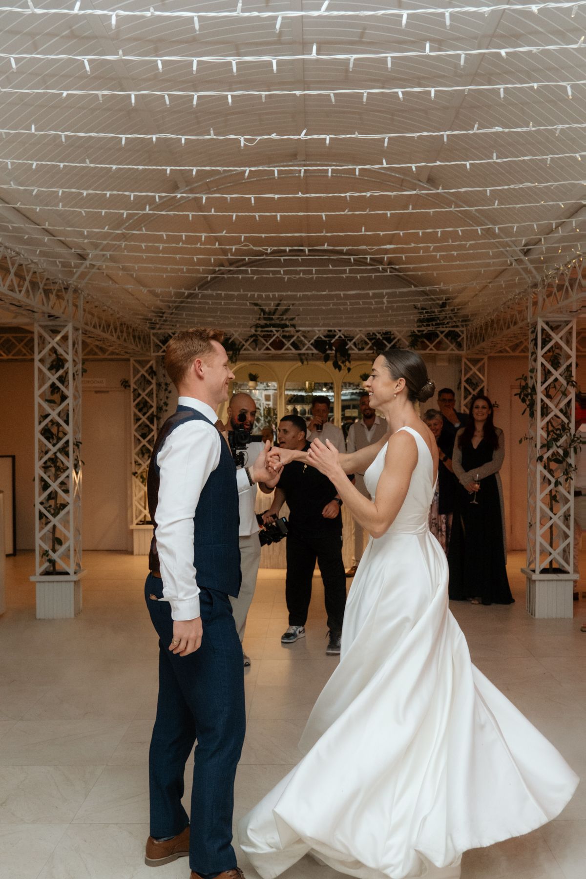 Bride in elegant white gown and groom dance joyfully under twinkling lights at wedding reception.