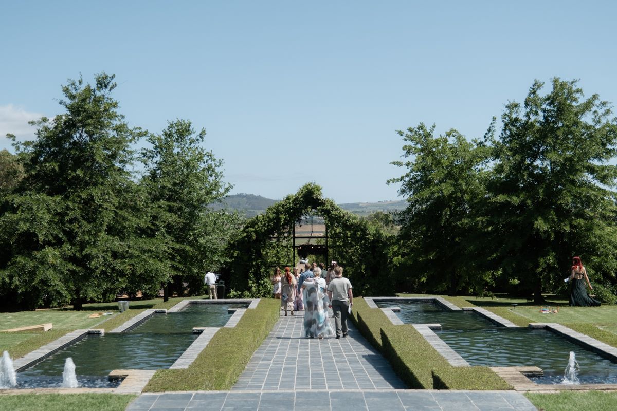 Wedding guests walk along a stone path flanked by reflective water features toward an ivy-covered ceremony arch.