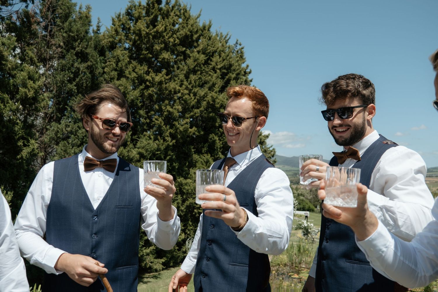 Three groomsmen in navy vests and sunglasses toasting with whiskey glasses outdoors on a sunny wedding day.
