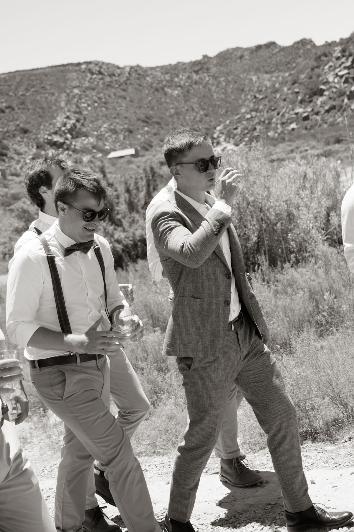 Two groomsmen in sunglasses walk outdoors at a desert wedding, one smoking and wearing a suit.