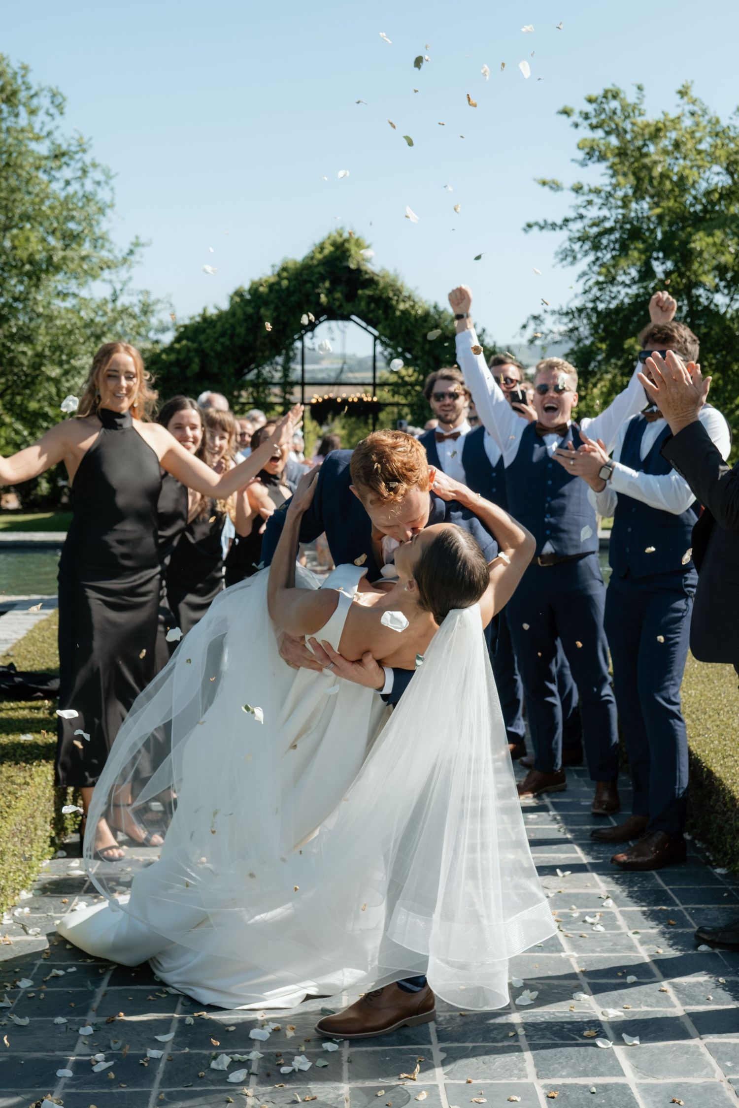 Bride and groom share a romantic dip kiss while wedding guests cheer and toss confetti outdoors.