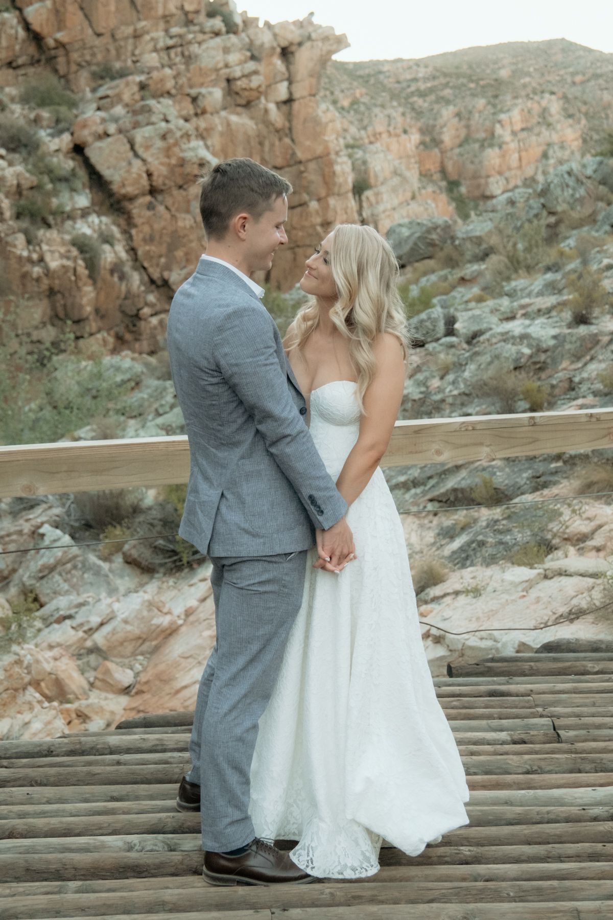 Bride in strapless lace gown and groom in grey suit holding hands on a wooden deck with rocky backdrop.