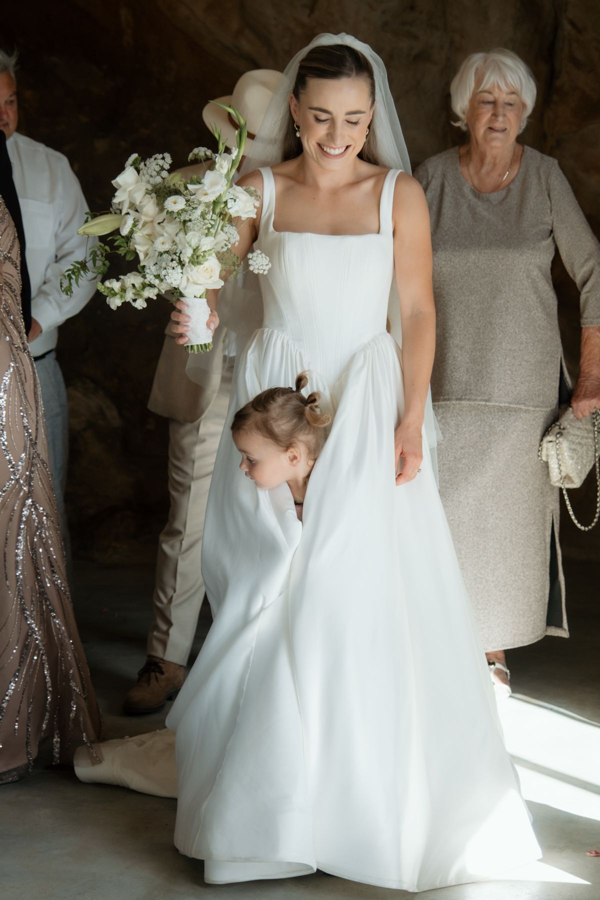 Smiling bride in white gown holding bouquet with flower girl adjusting her train at wedding ceremony.