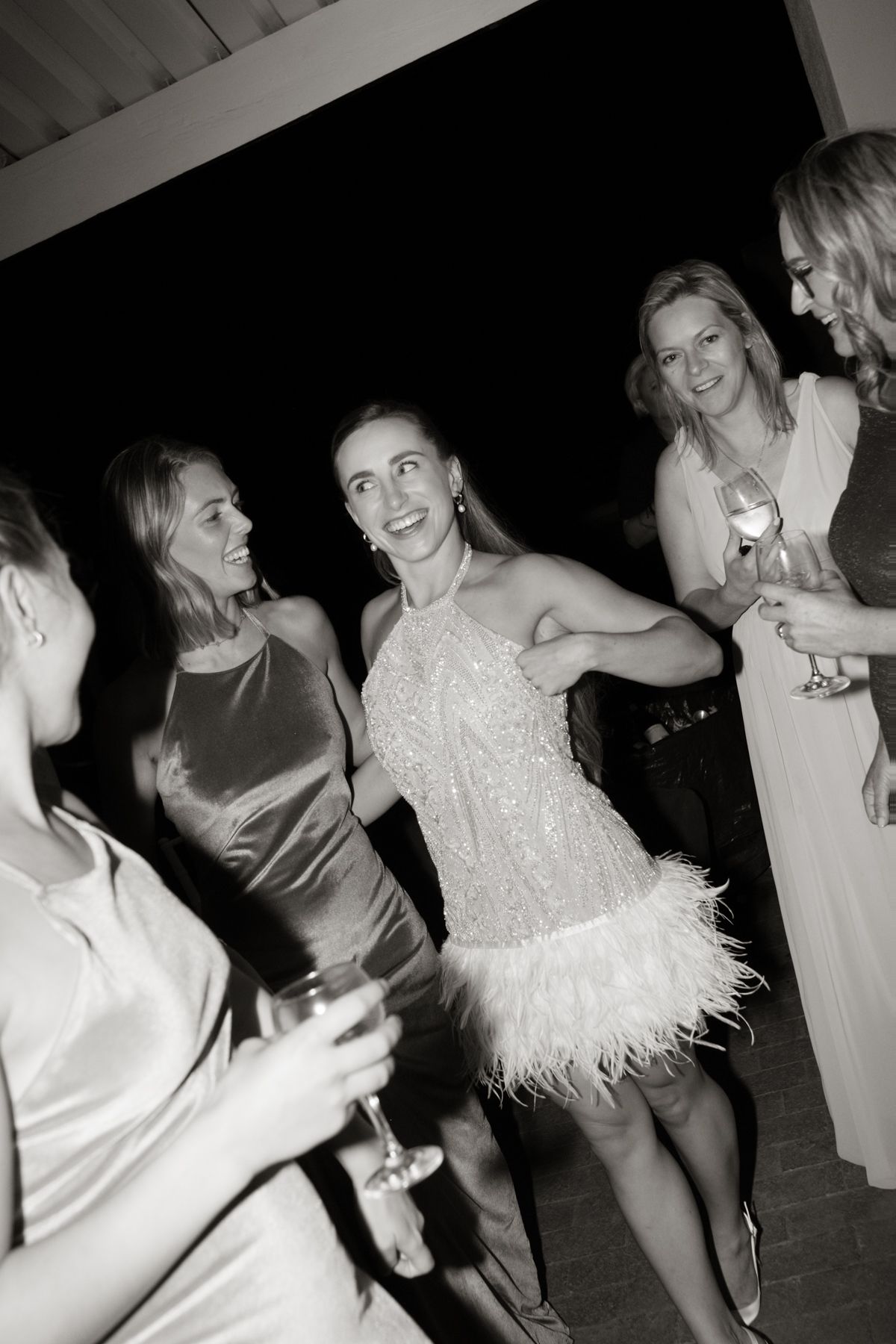 Black and white photo of bride in feather dress dancing with guests at wedding.
