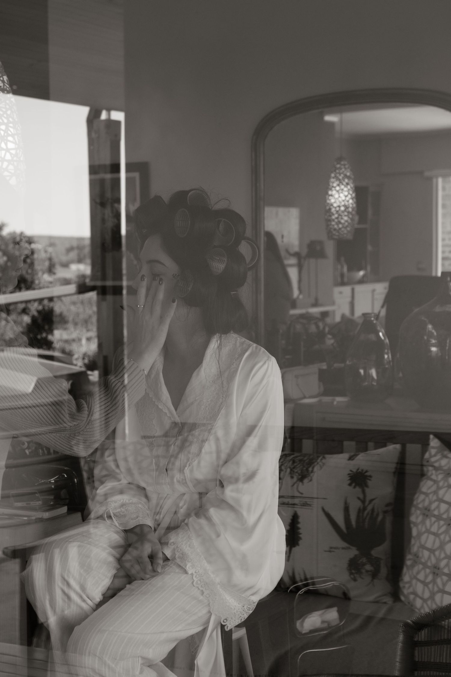 Black and white photo of a bride in hair rollers applying makeup while seated, reflected in a window.