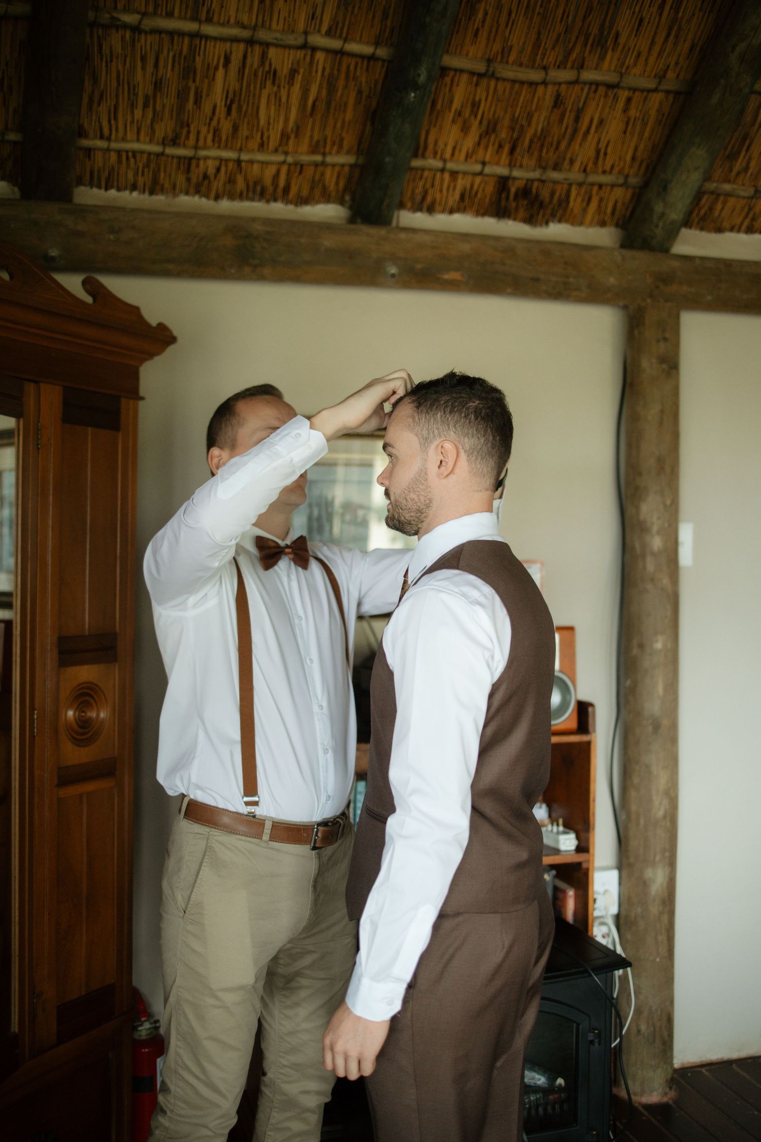Two groomsmen in white shirts and suspenders help each other prepare for a wedding ceremony indoors.
