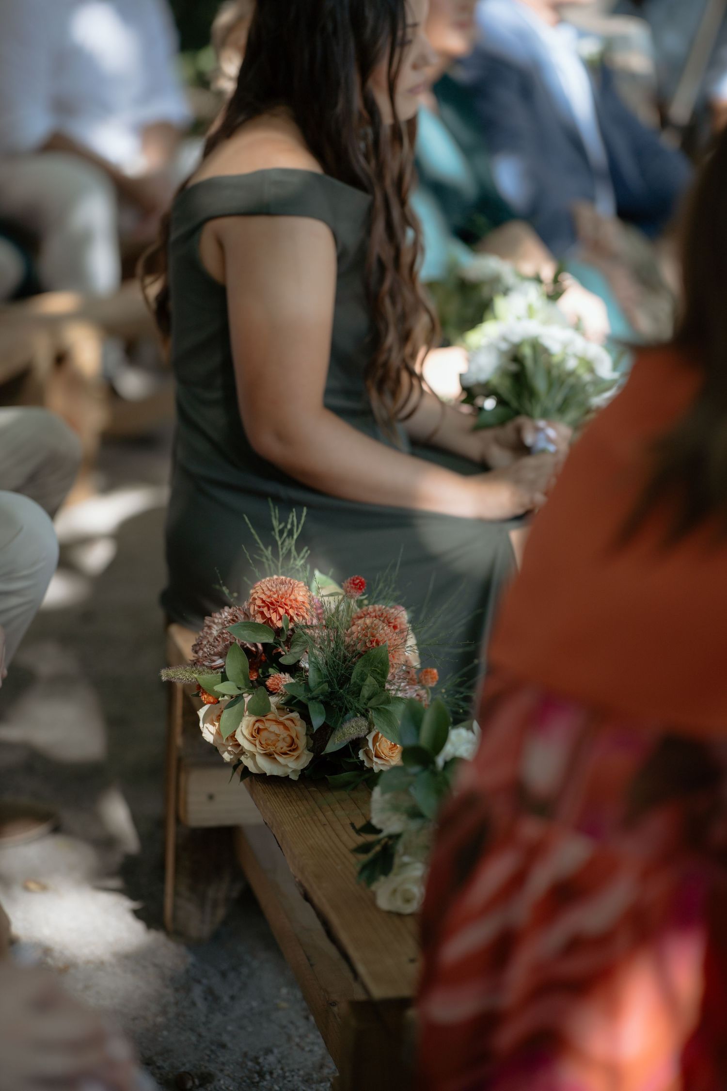 A bridesmaid in an off-shoulder sage green dress sits on a wooden bench beside a bouquet of peach and white flowers.