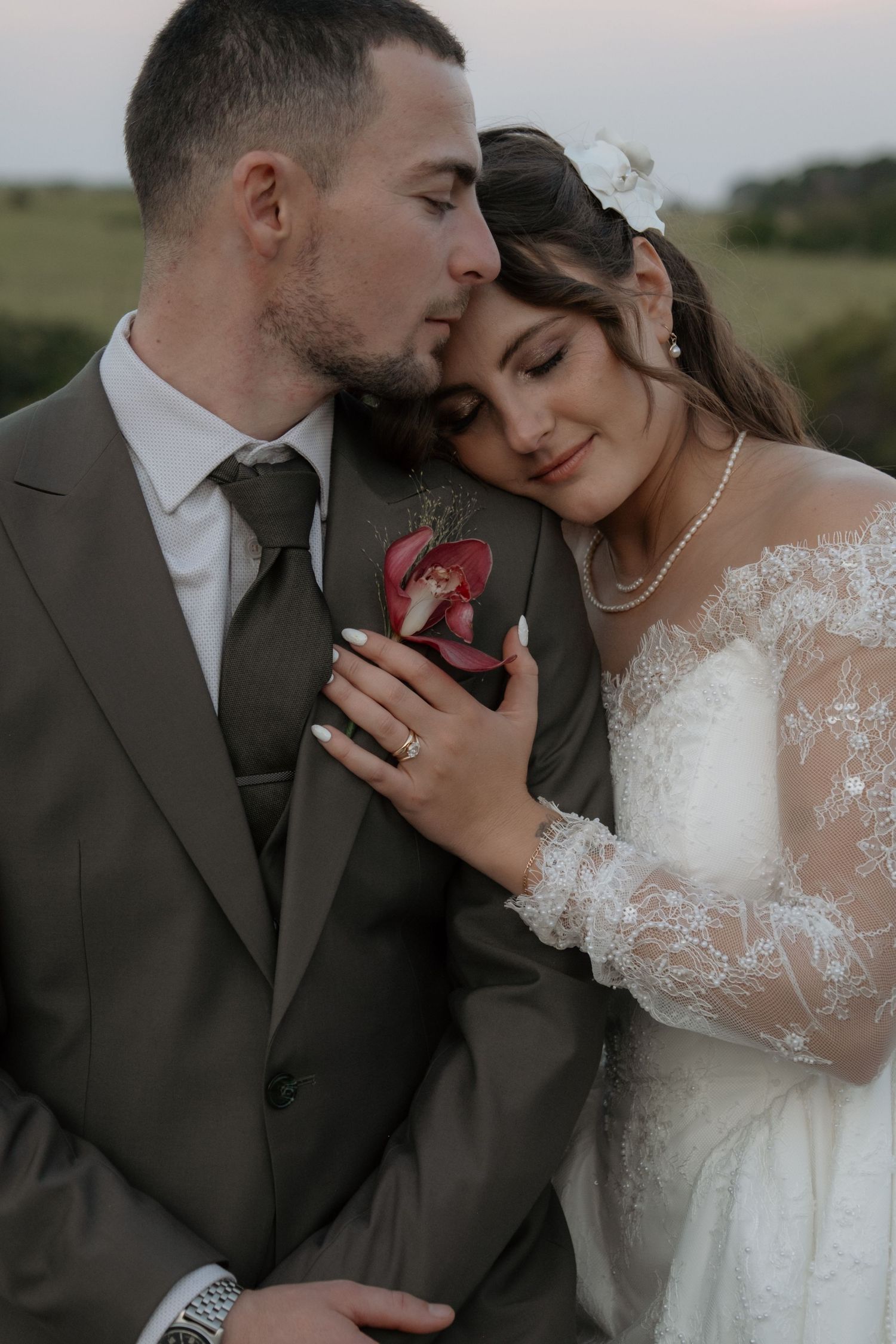 Romantic wedding couple embracing at dusk, bride in lace gown resting head on grooms gray suit.