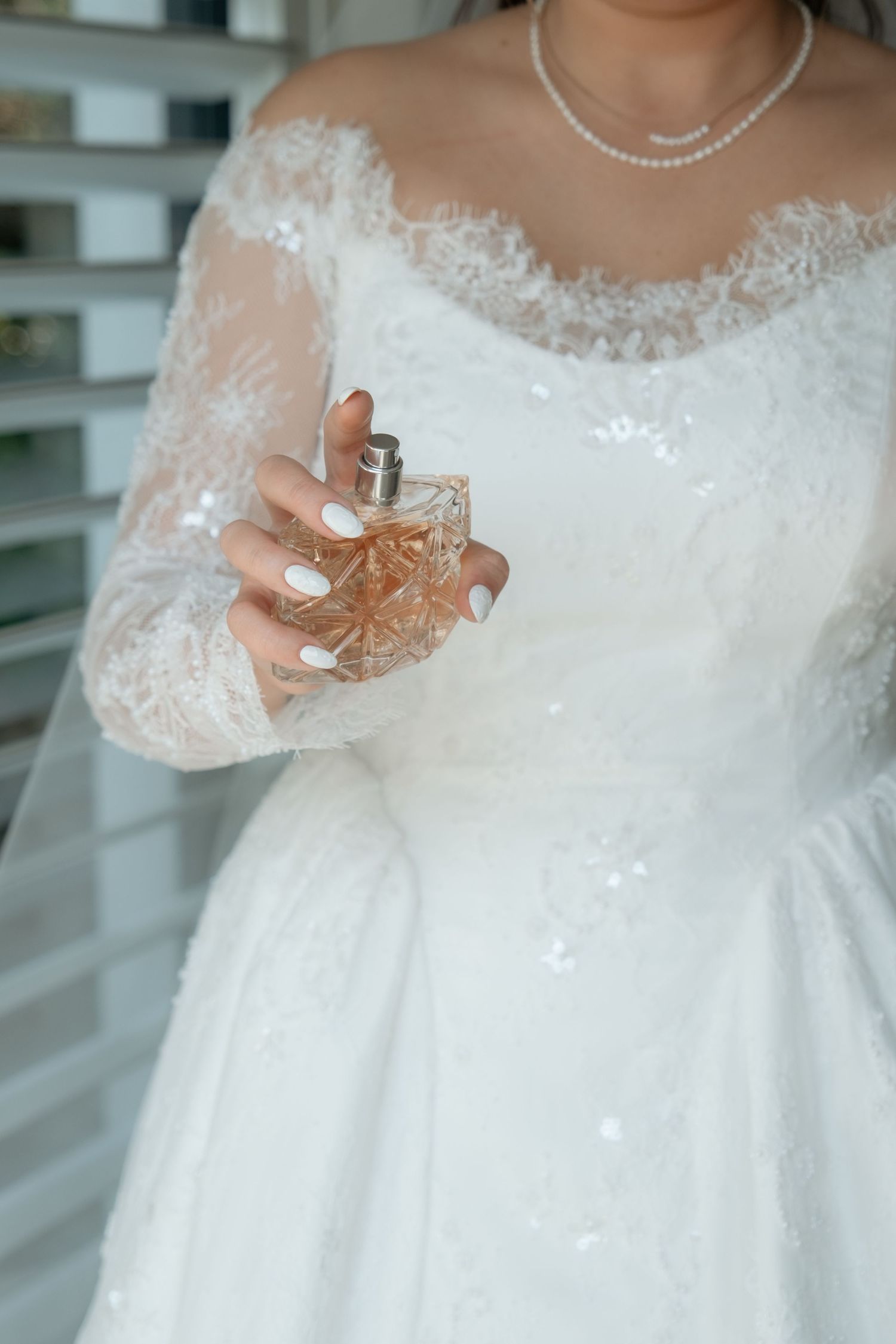 Bride in lace off-shoulder wedding gown holding a crystal perfume bottle with white manicured nails.