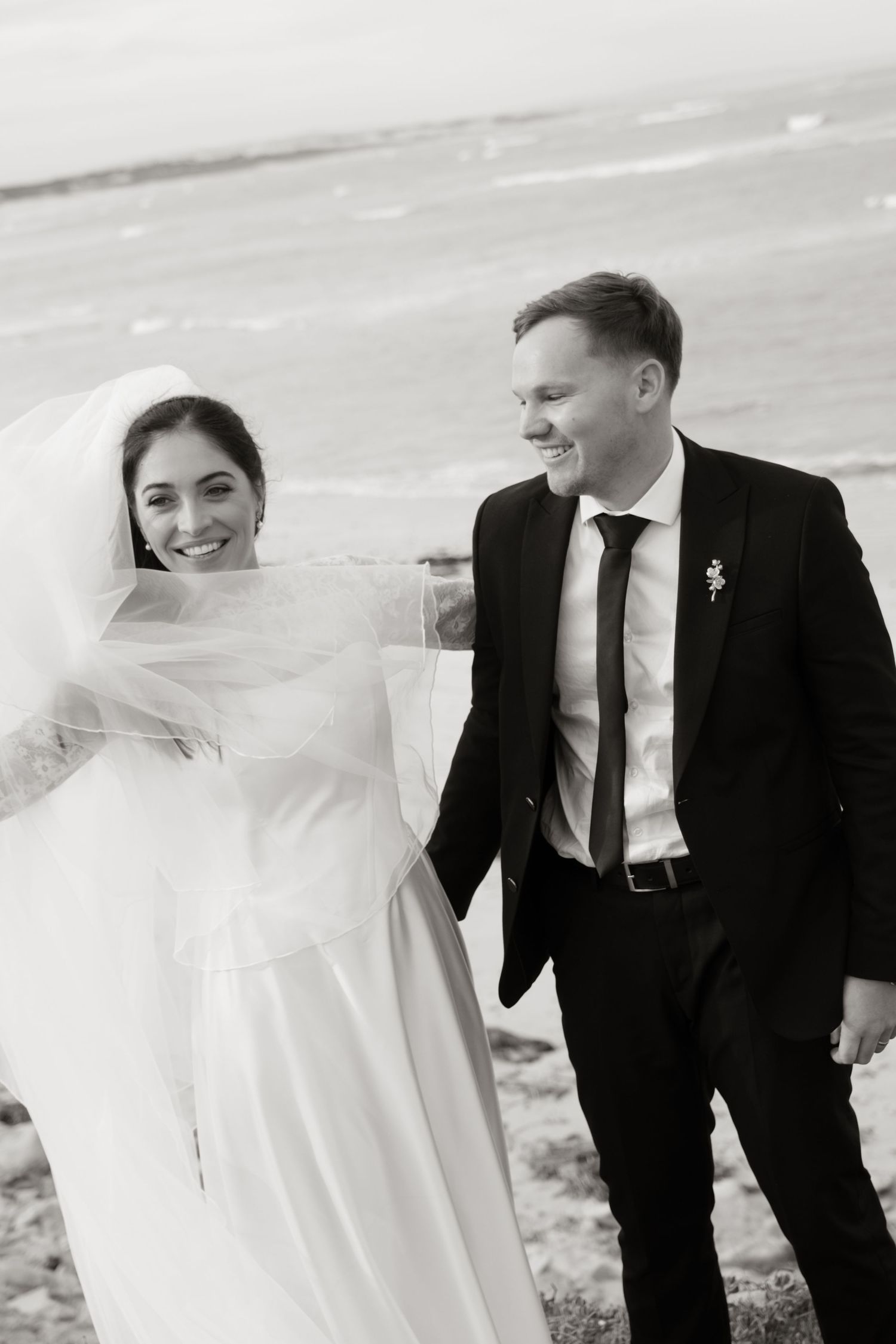 Black and white photo of smiling bride and groom walking by the ocean on their wedding day.
