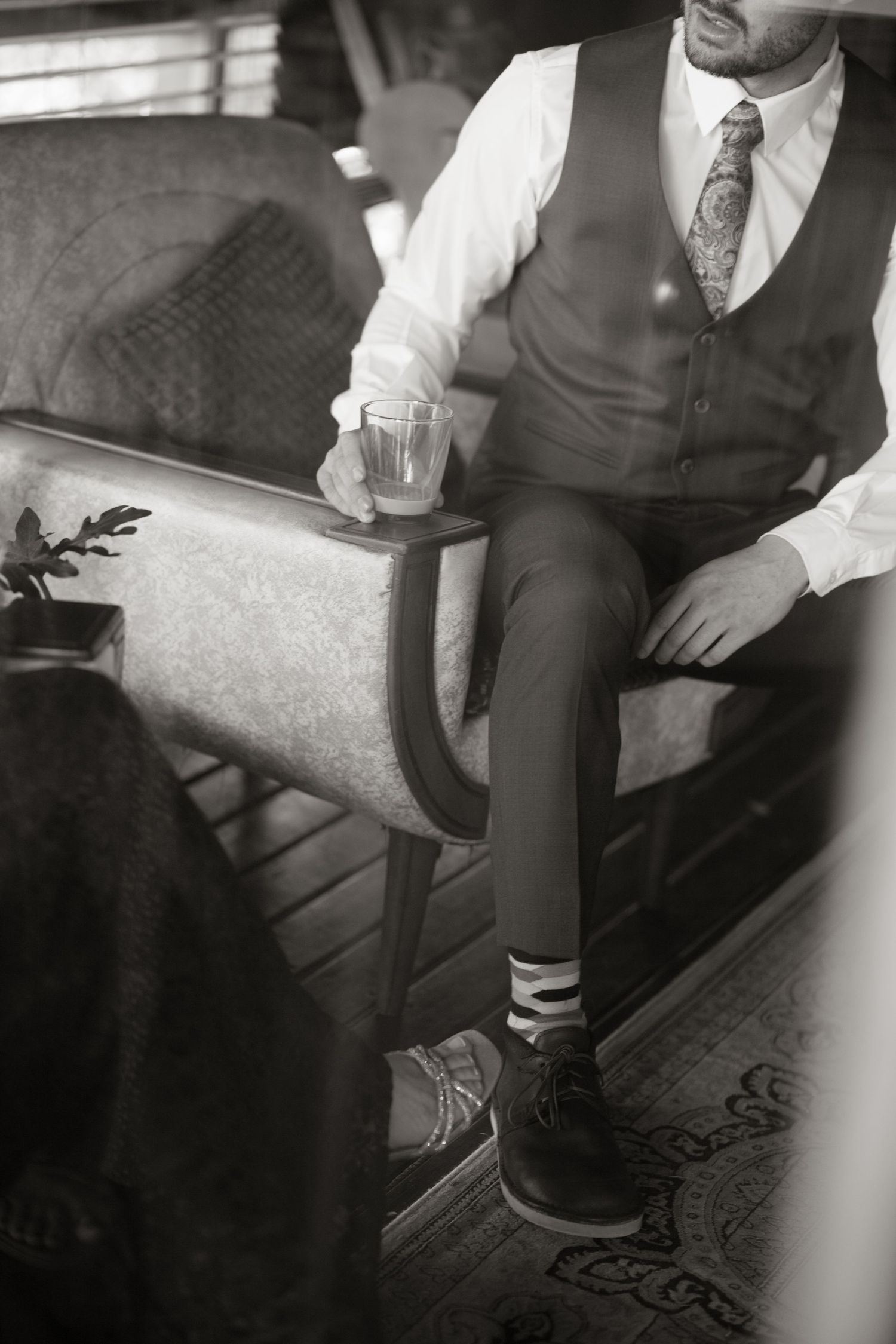 Groom in brown vest and tie sits in vintage chair holding glass of whiskey, black and white wedding photo.