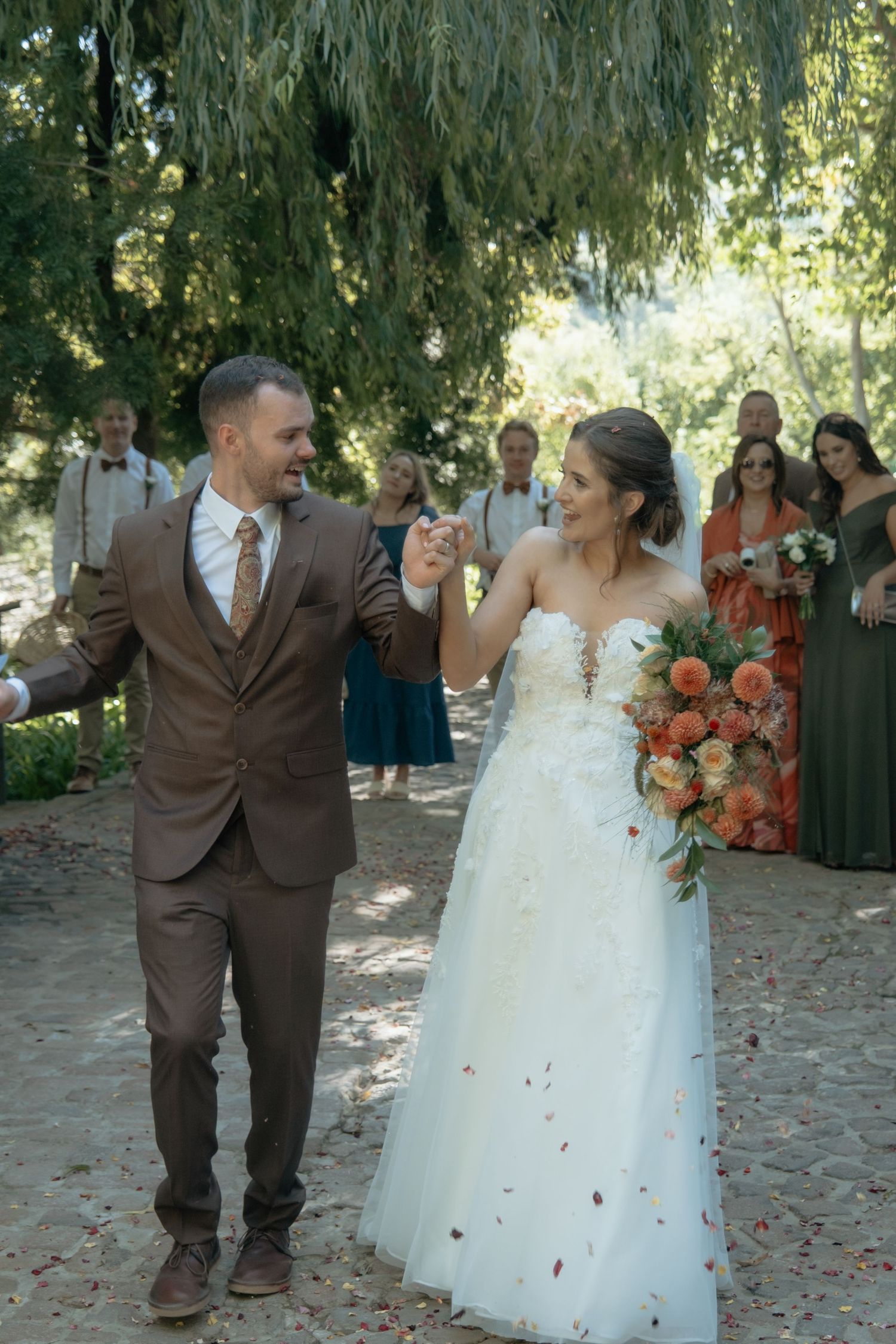 Newlyweds walking hand in hand down outdoor aisle as guests celebrate with confetti in lush garden setting.