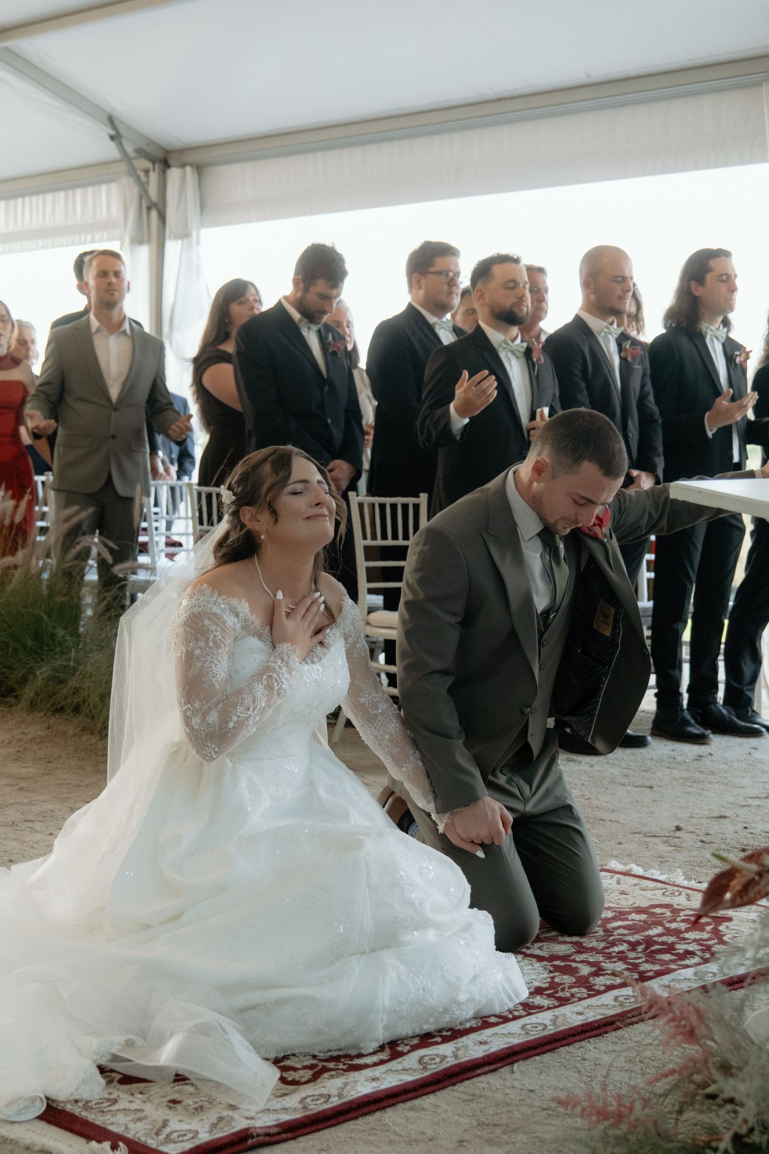 Bride and groom kneeling in prayer during outdoor wedding ceremony, guests standing behind them under white tent.