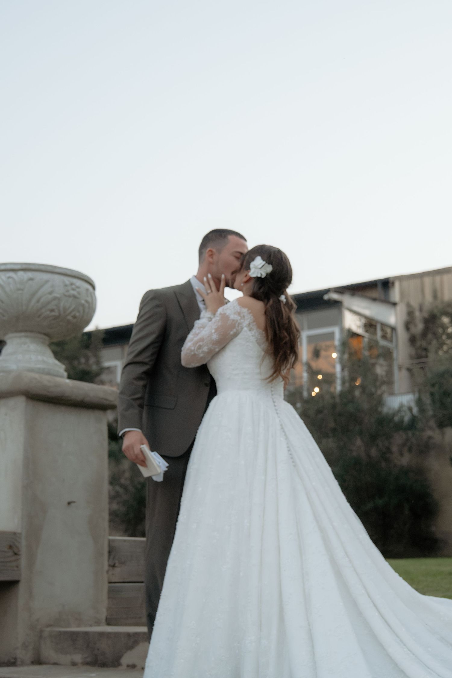 Bride and groom sharing a romantic kiss outdoors at dusk near stone urn.