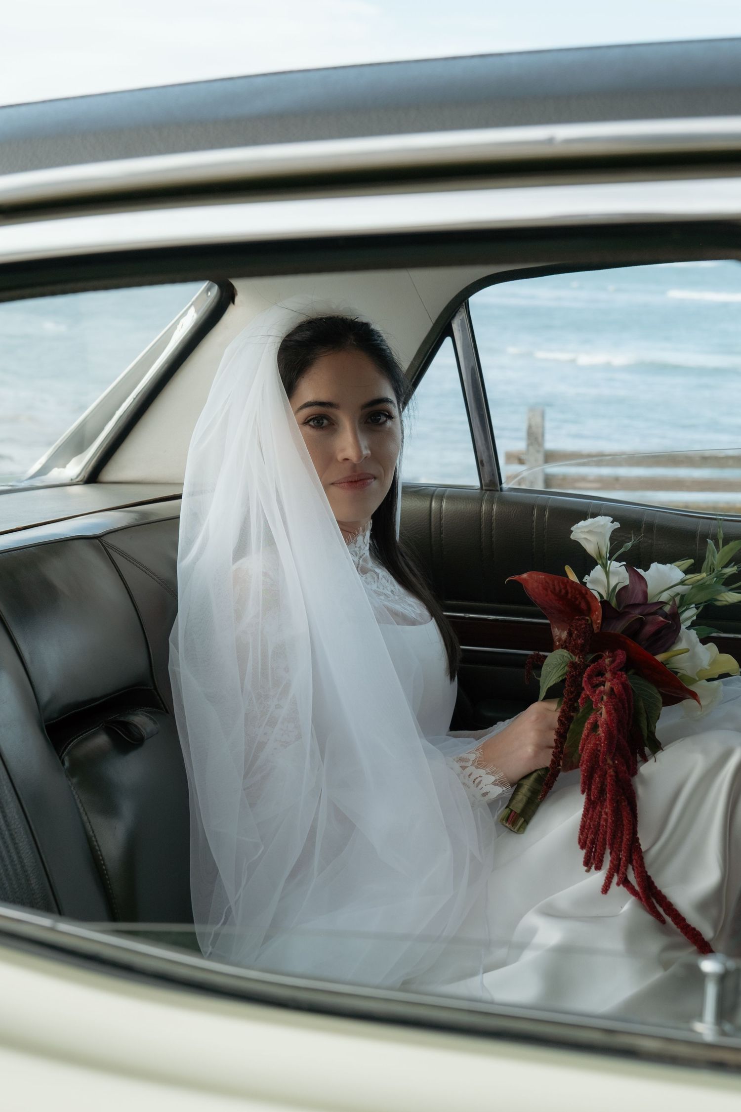 Elegant bride in white gown and veil sits in vintage car holding red and white bouquet by the ocean.