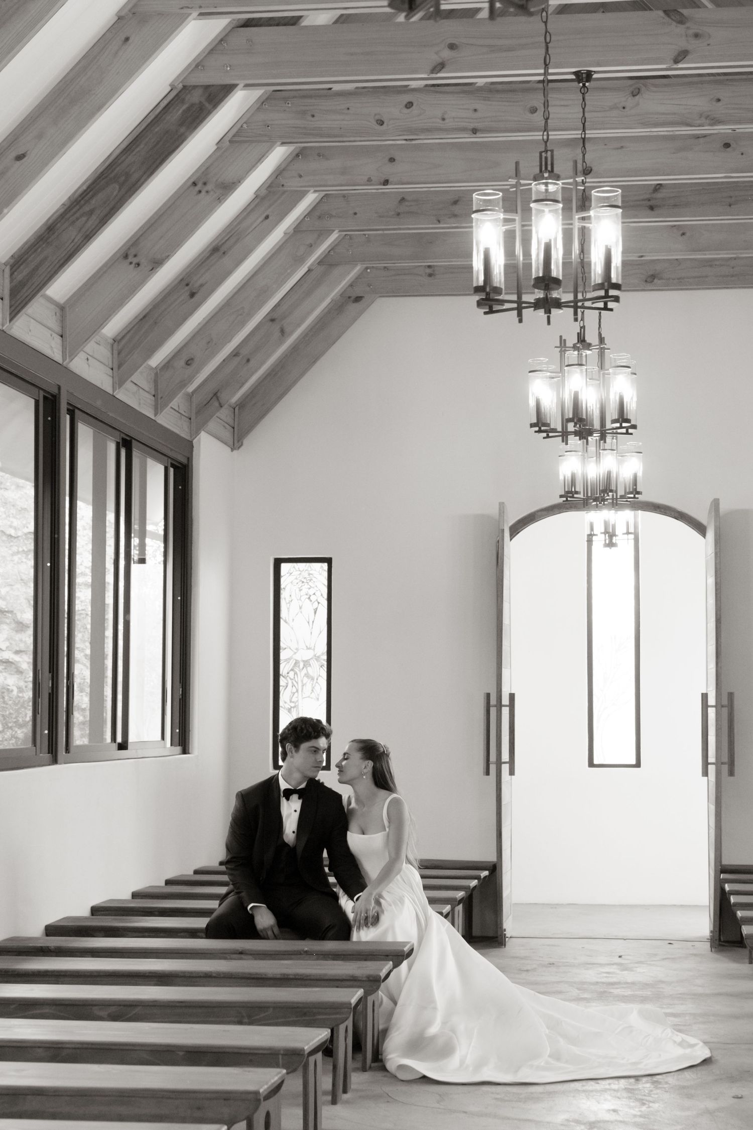 A couple shares an intimate moment in a bright white chapel with arched windows and wooden pews.