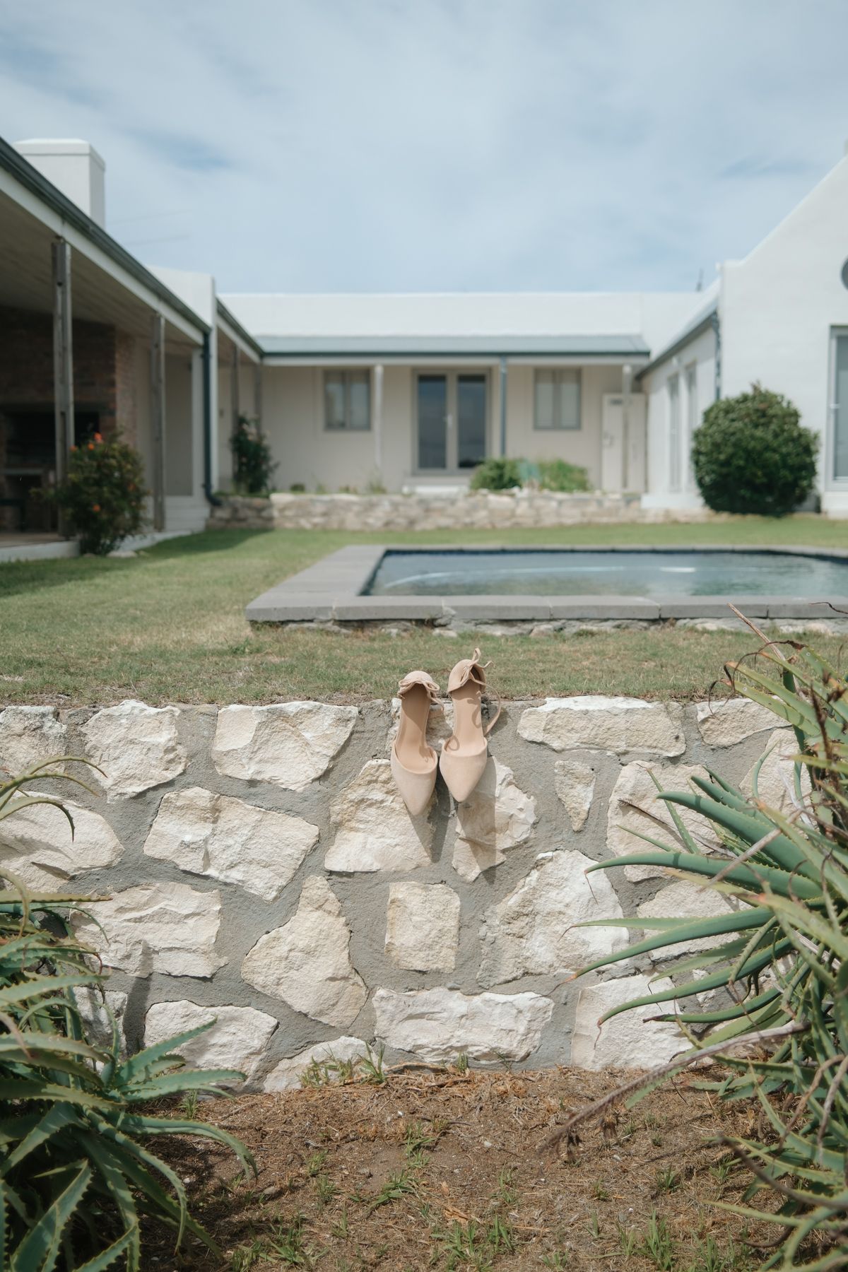 A stone retaining wall extends along the front of a white residential building with a lawn and cloudy sky.