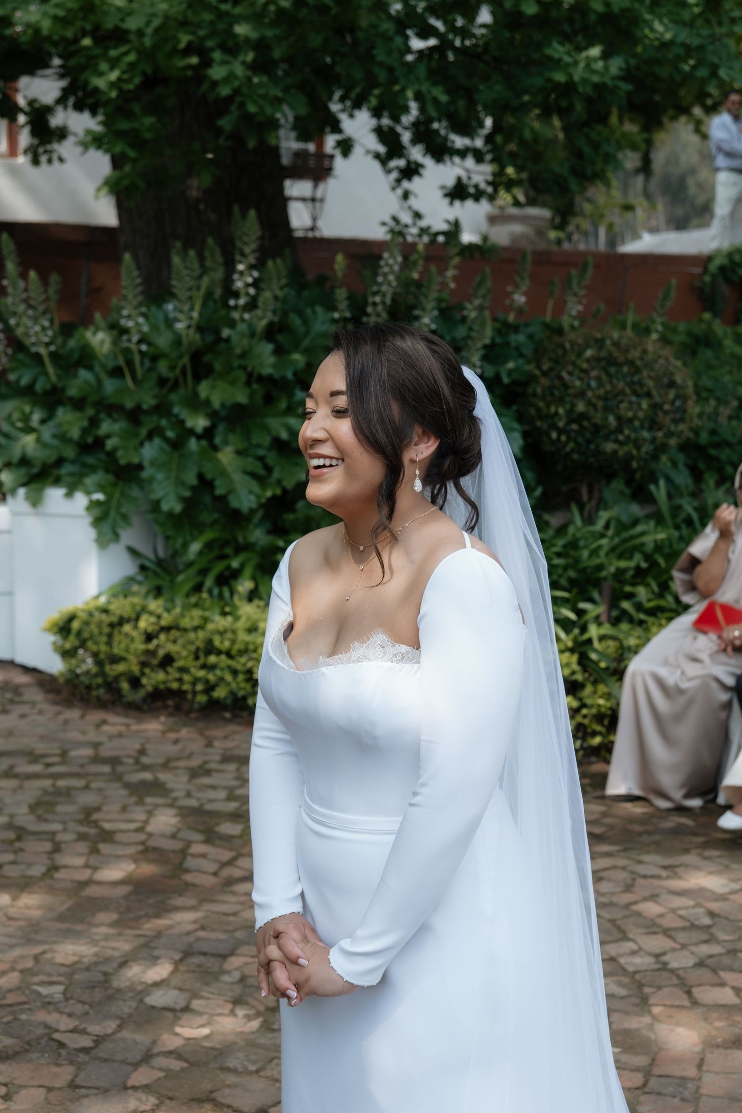 Smiling Asian bride in a fitted long-sleeve white wedding gown and veil standing outdoors among lush greenery.