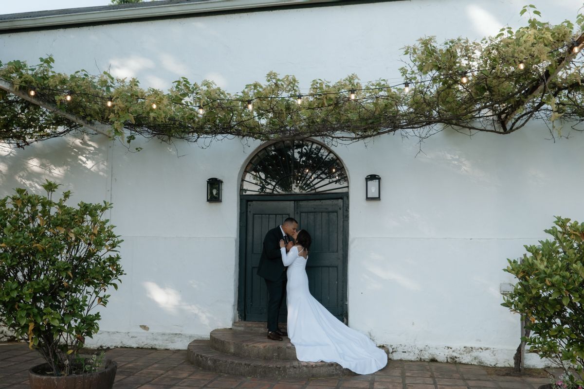 Bride and groom sharing a kiss in front of a white building with arched green door.