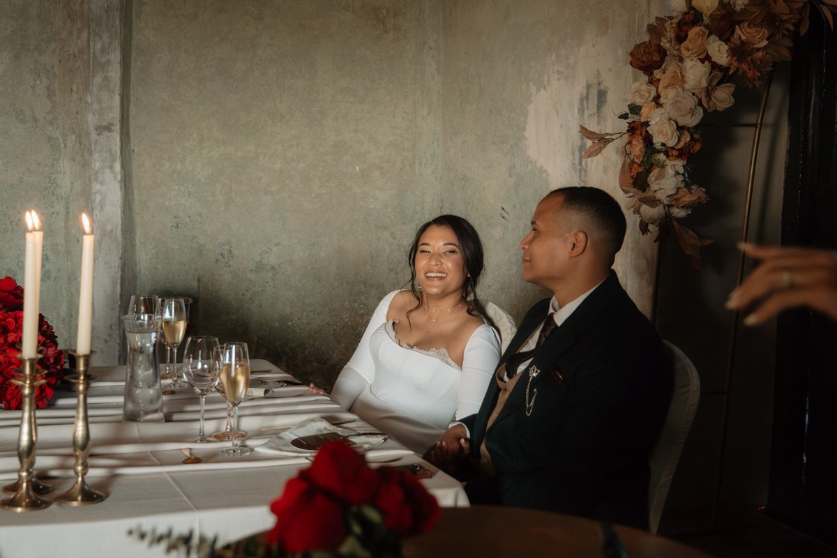 Bride in white strapless gown laughs joyfully beside groom in black suit at elegant candlelit wedding sweetheart table.