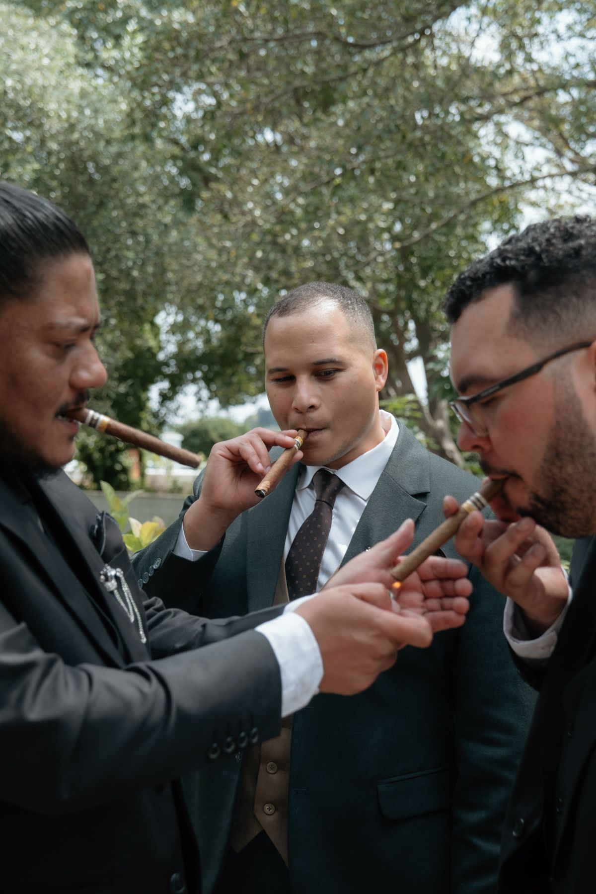 Three groomsmen in dark suits smoking cigars outdoors before a wedding ceremony.