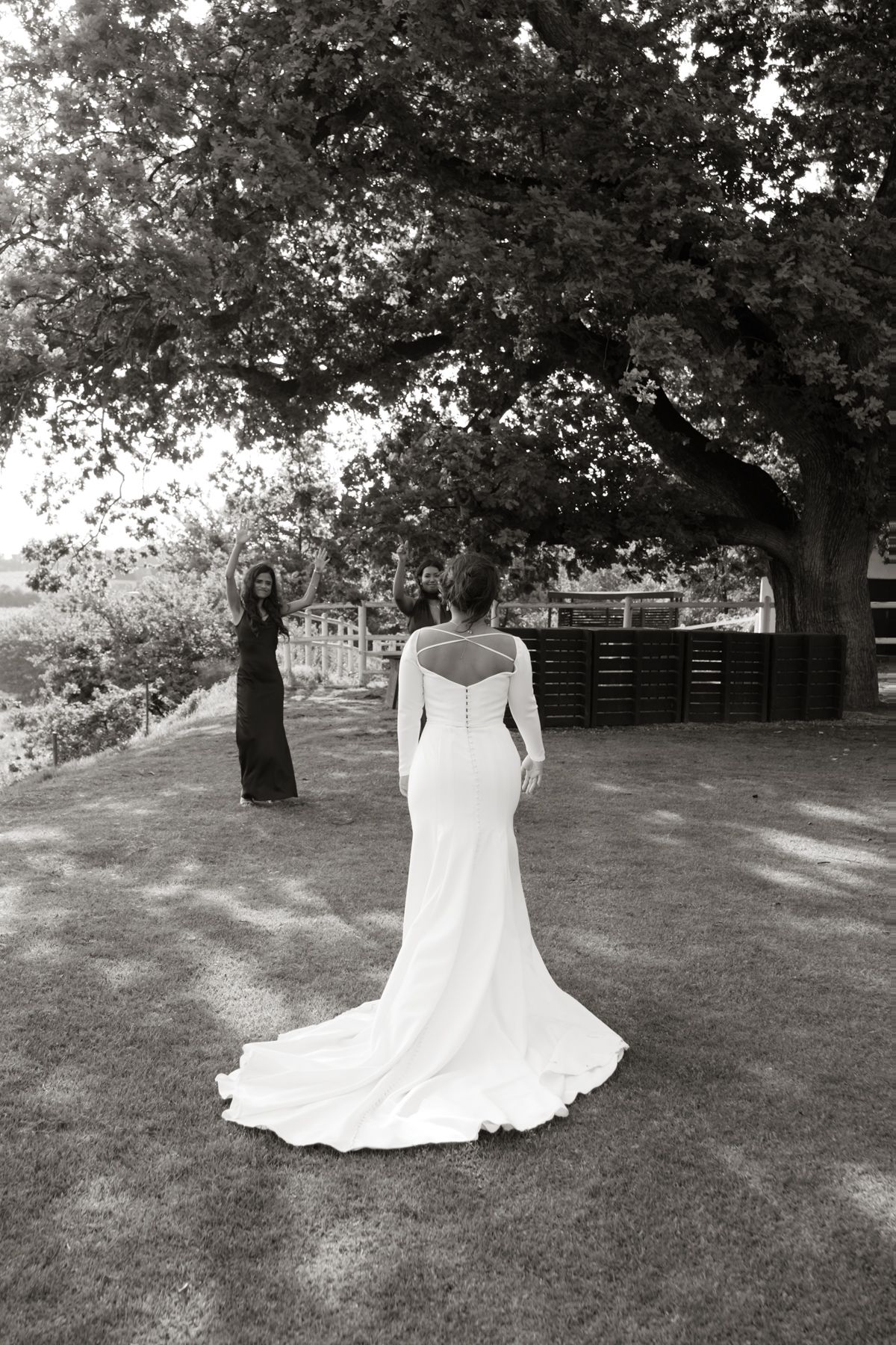 Black and white photo of bride in long-sleeved gown with train as bridesmaids react with excitement outdoors.