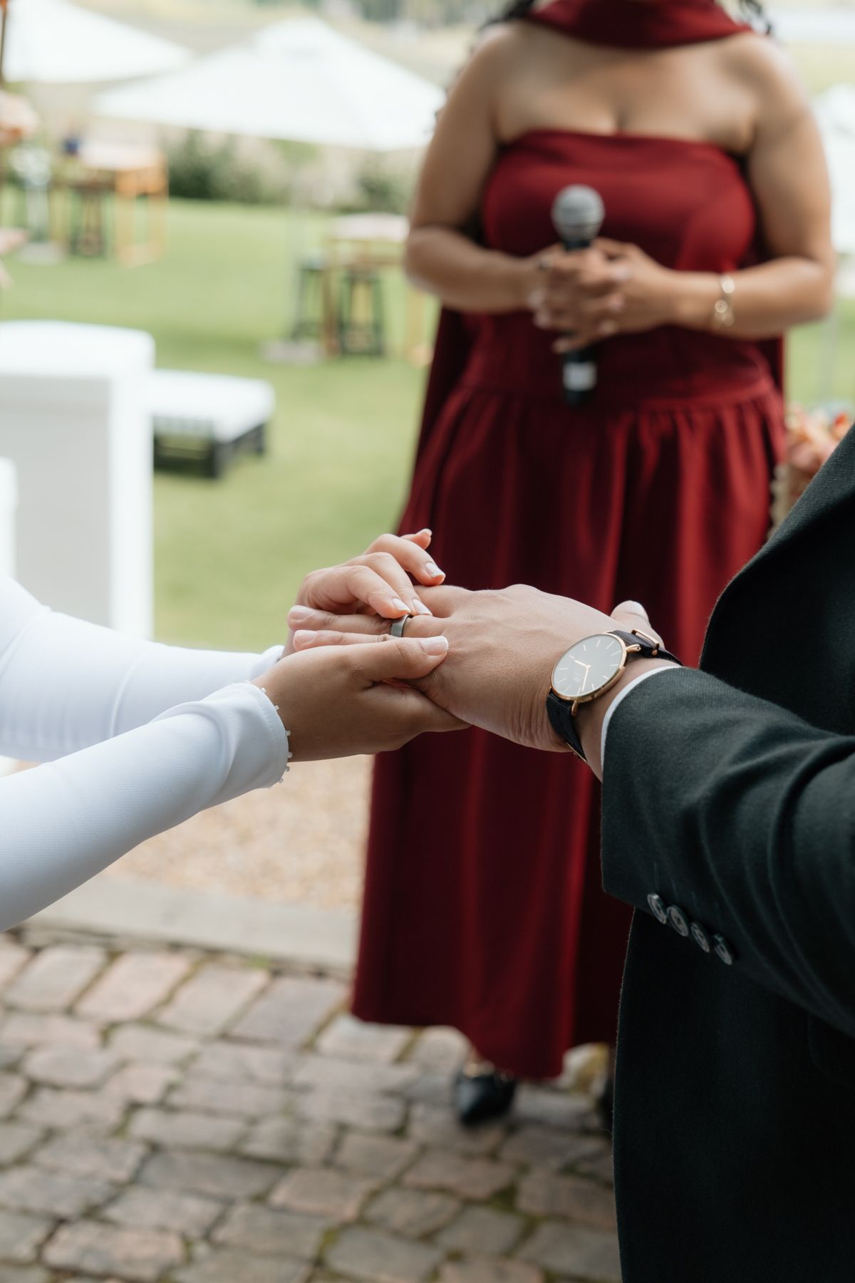 Groom placing wedding ring on bride's finger during outdoor ceremony while officiant in red dress holds microphone.