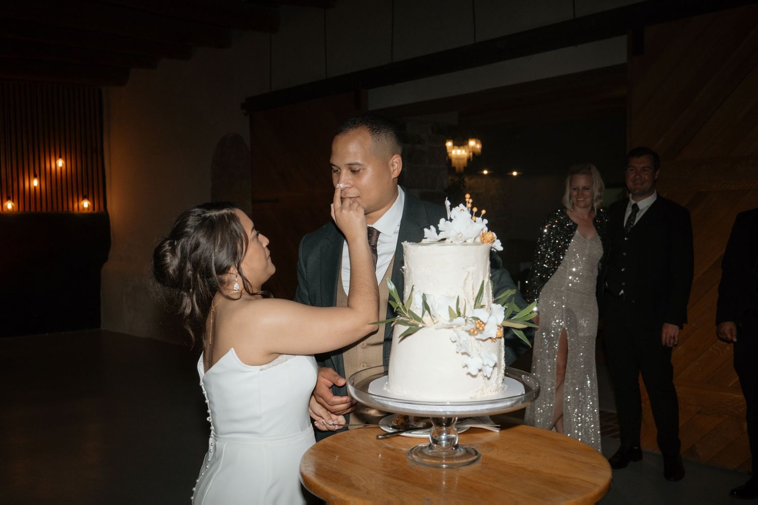 Bride playfully feeds wedding cake to groom at rustic reception venue while guests look on smiling.
