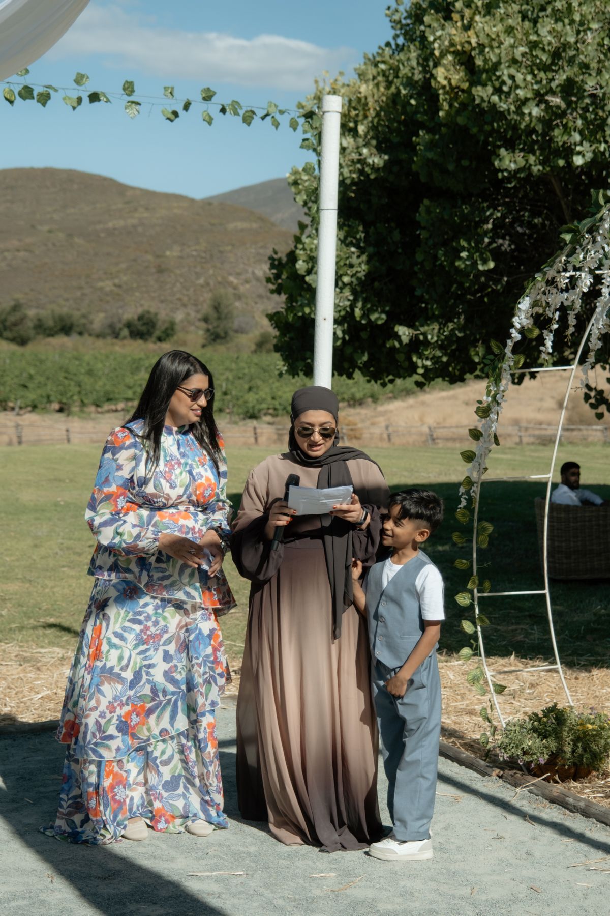 Two women and a child reading from papers during outdoor wedding ceremony with mountains behind.