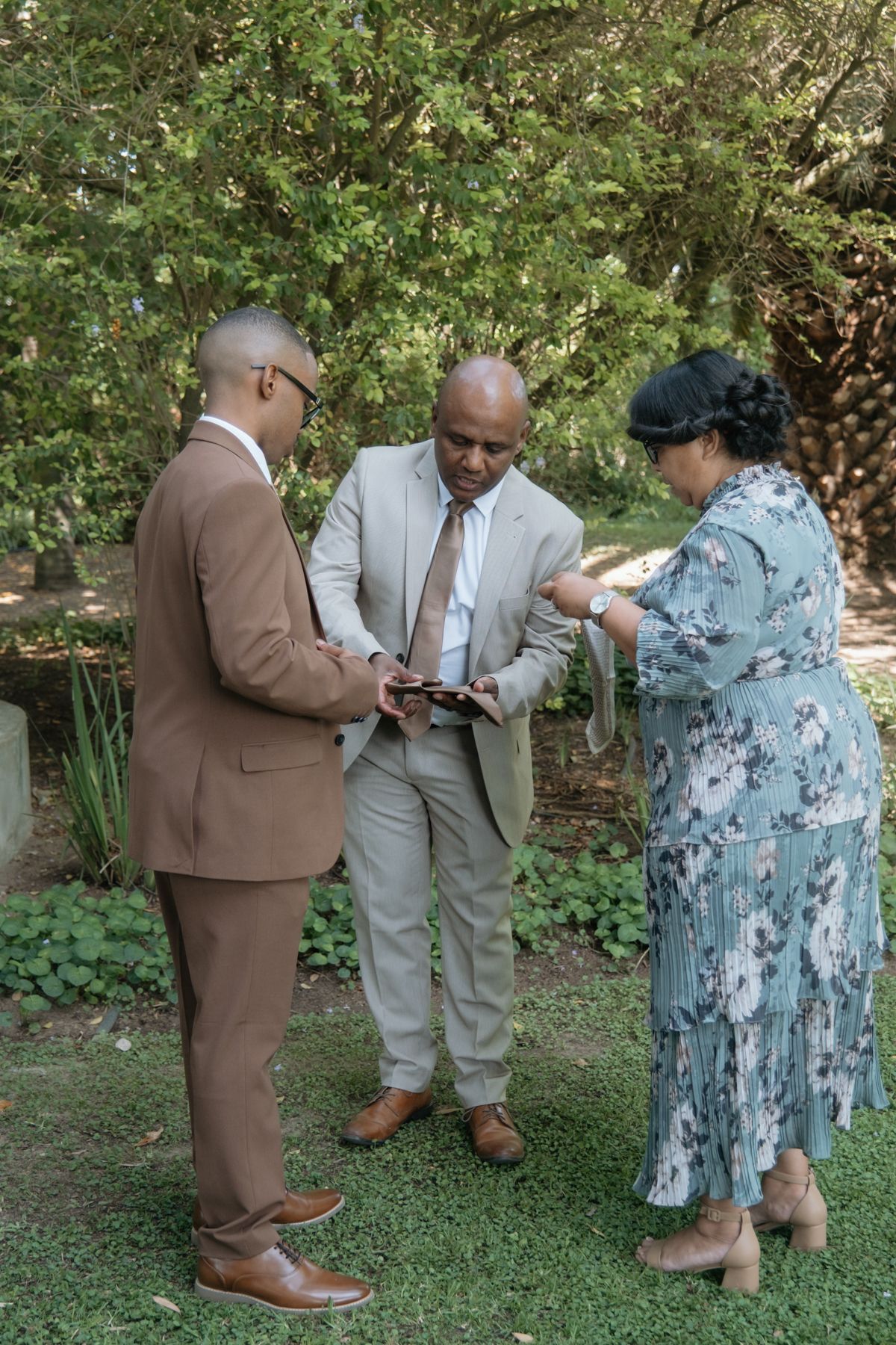 Two men in suits and a woman in a floral dress examine an item together in a garden setting.