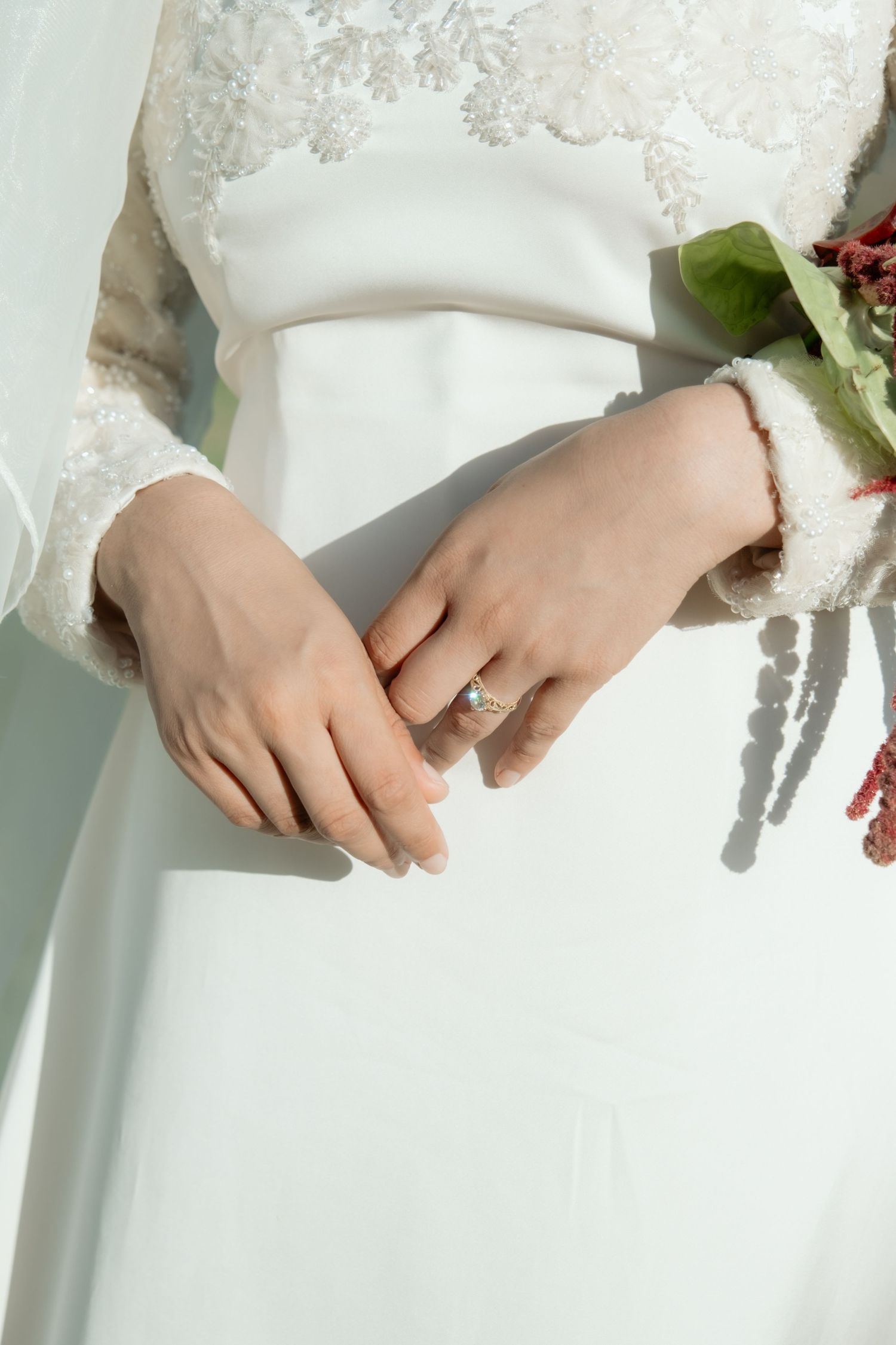 Close-up of bride's hands wearing elegant diamond engagement ring, holding bouquet in white lace wedding dress.