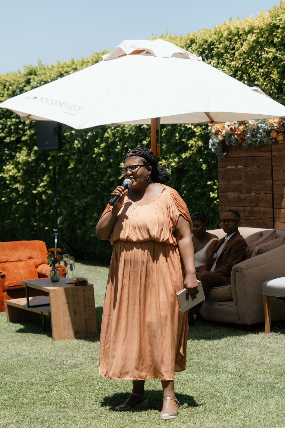 Woman in terracotta dress giving speech at outdoor wedding reception under white umbrella.