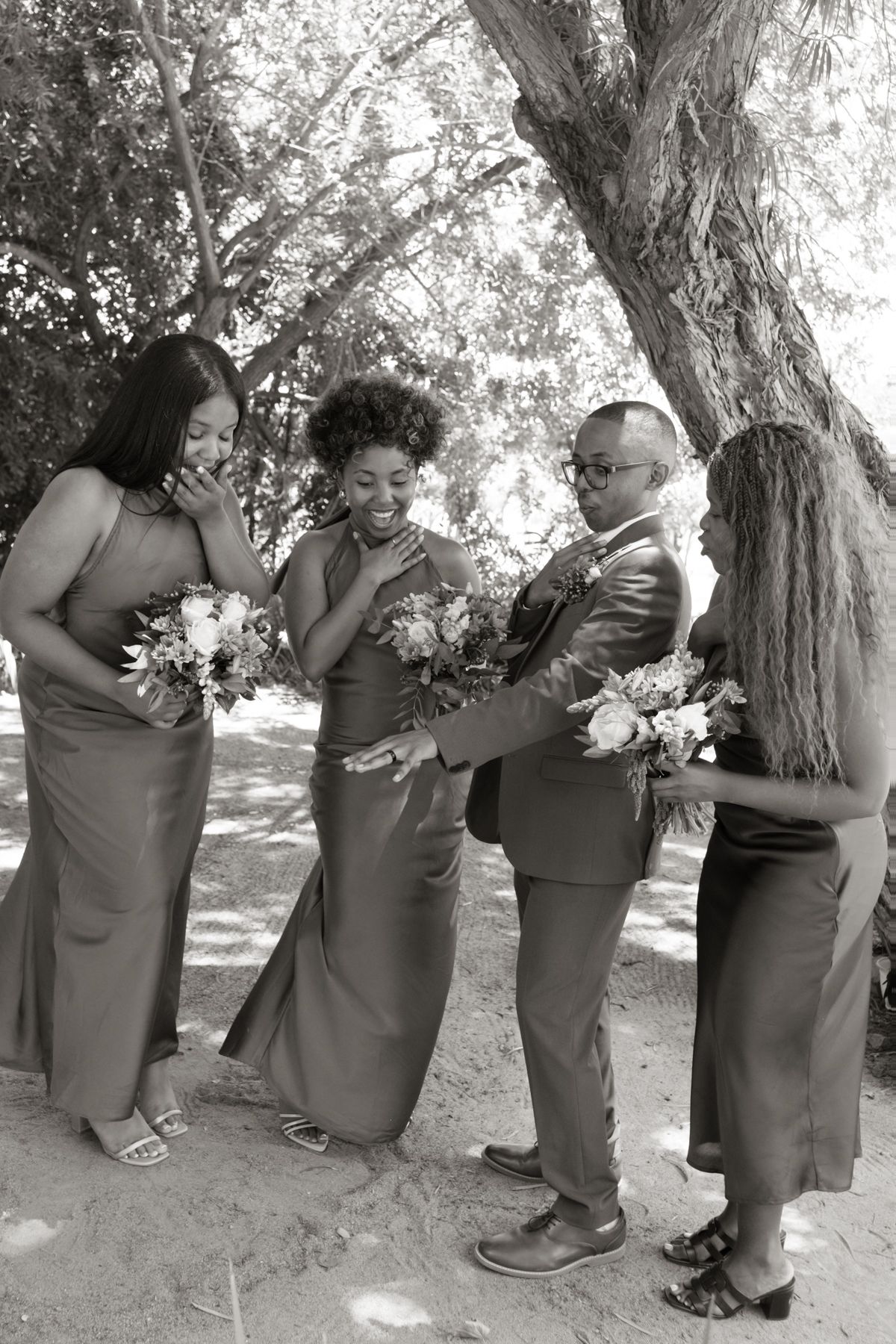 Black and white photo of bride, groom, and bridesmaids laughing together while holding bouquets at an outdoor wedding.