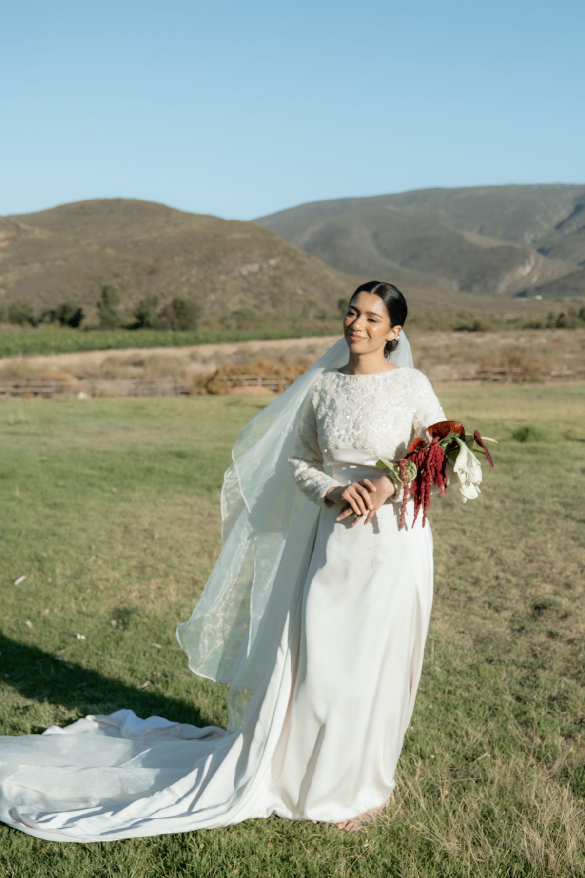 Smiling bride in white lace gown and veil holds red bouquet in scenic mountain landscape.