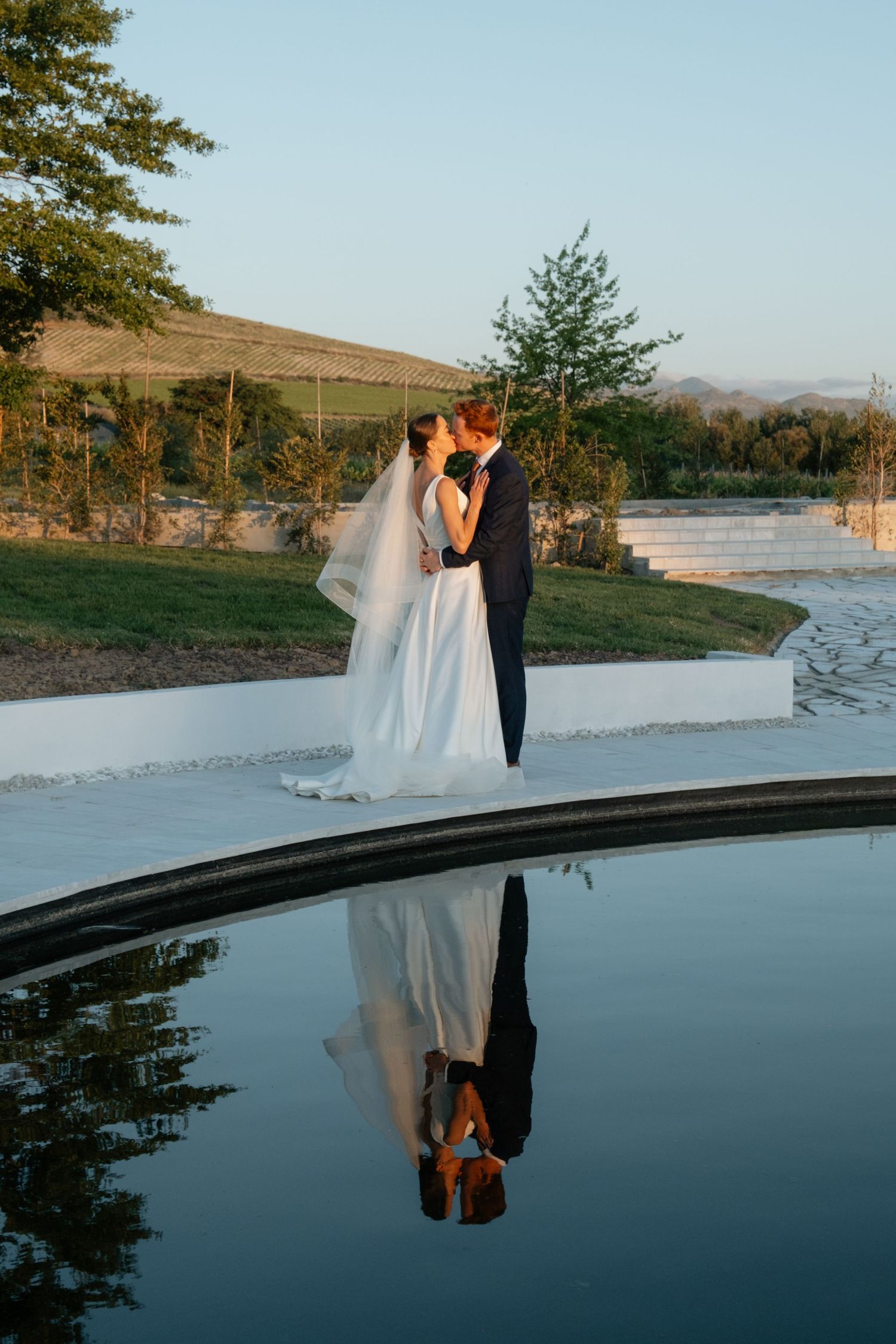 Bride and groom share a romantic kiss beside a reflective pool, with their mirror image visible in the still water.